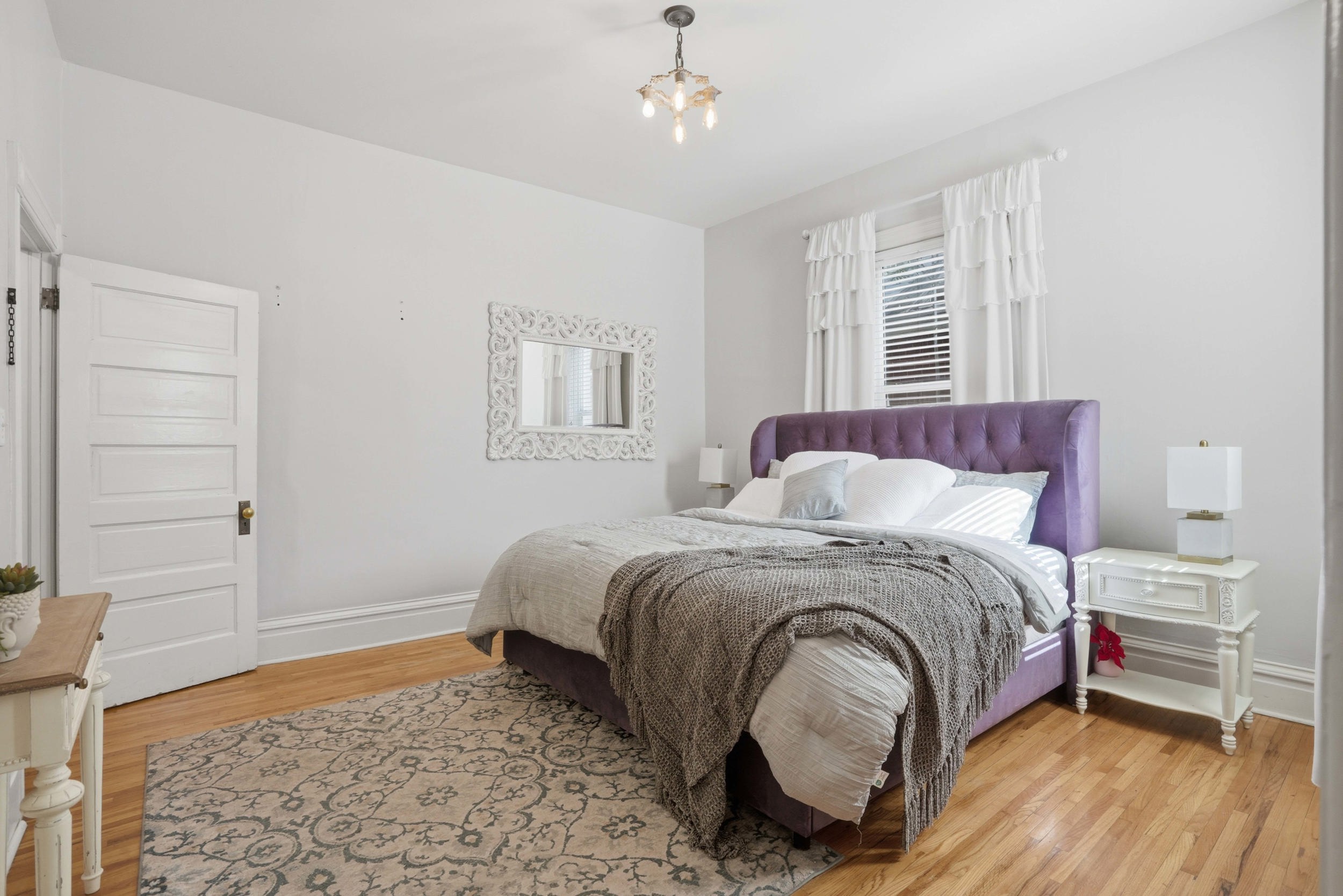 Bedroom featuring wood-finish flooring, white trim, and a window with blinds and ruffled white curtains