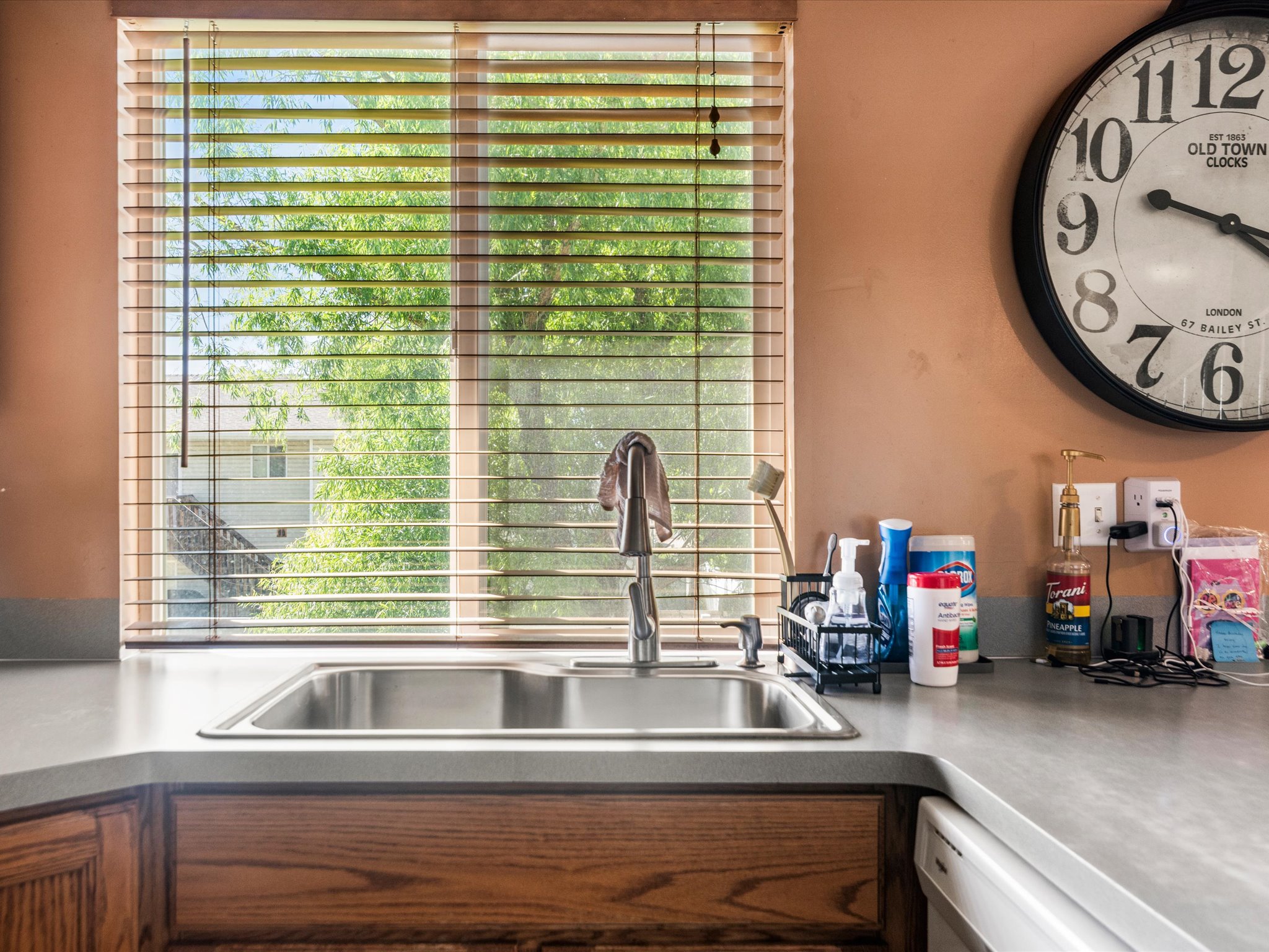 Kitchen featuring a stainless steel double-basin sink, a chrome pull-down faucet, wood-finish cabinetry, light-toned countertops, and a window with horizontal blinds