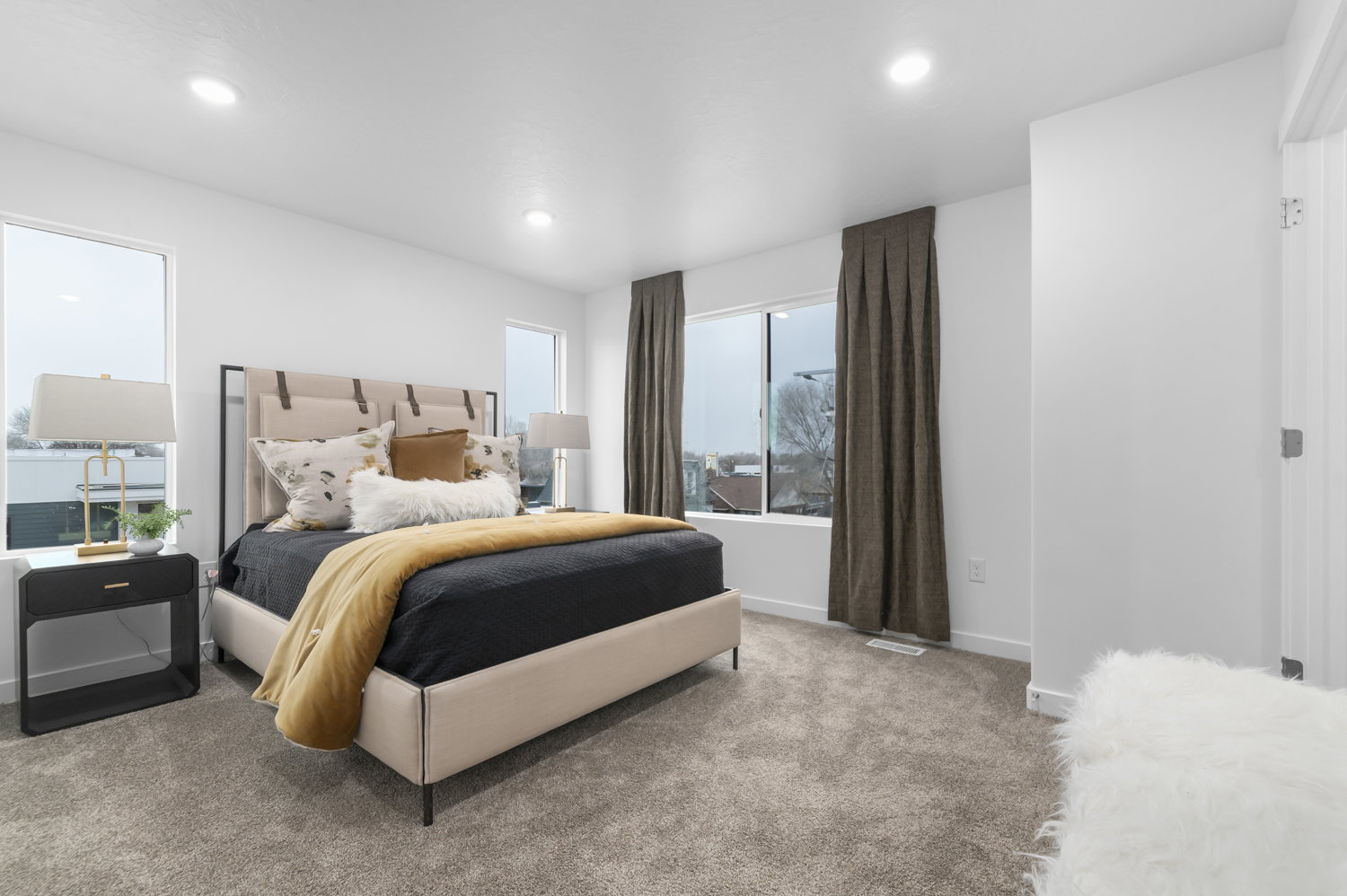 Spacious bedroom featuring light neutral wall paint, recessed lighting, expansive windows with dark drapery, and soft grey carpeting