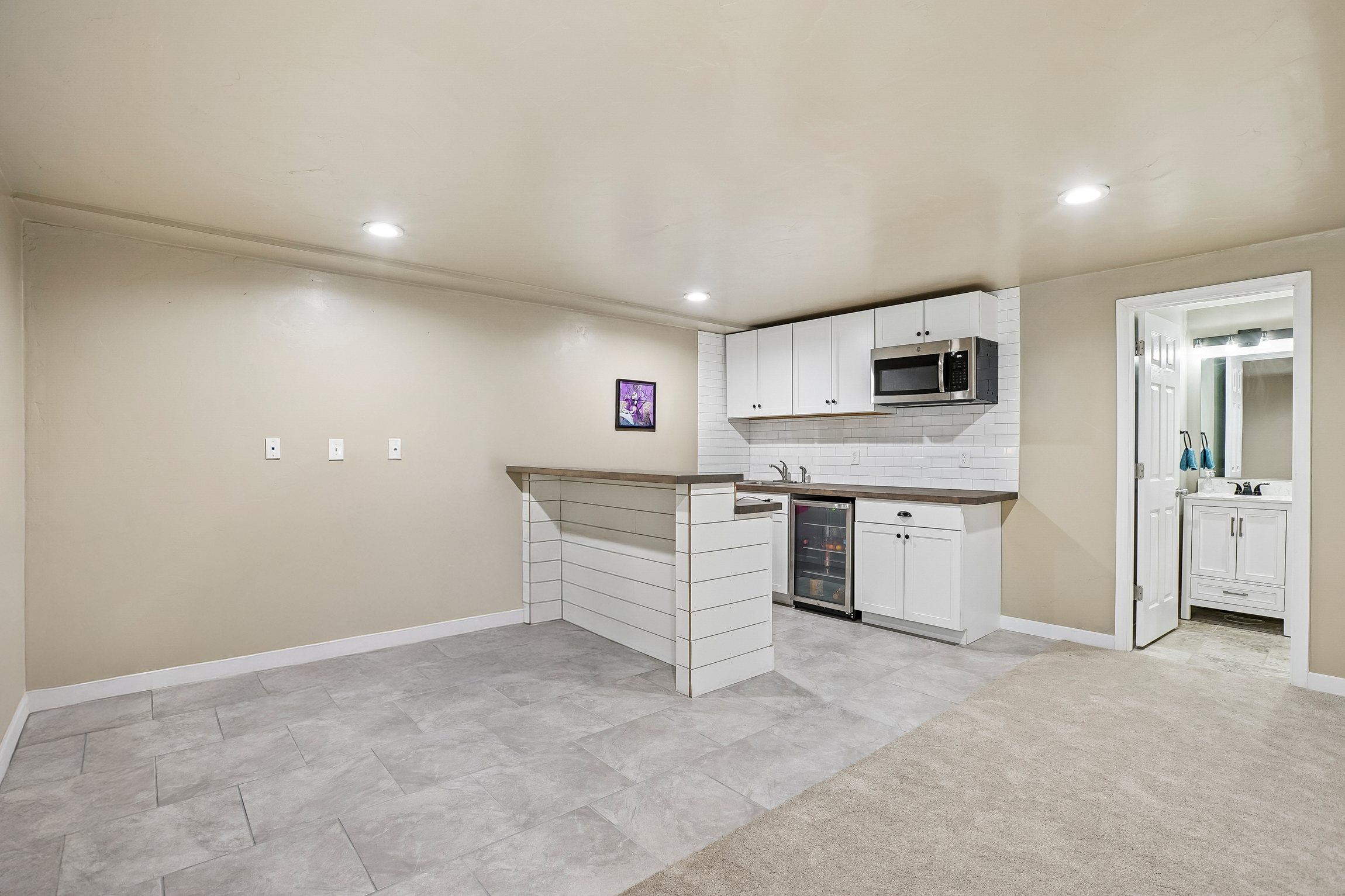 Open-concept living space featuring a wet bar, stainless steel microwave, beverage refrigerator, white cabinetry, and a subway tile backsplash