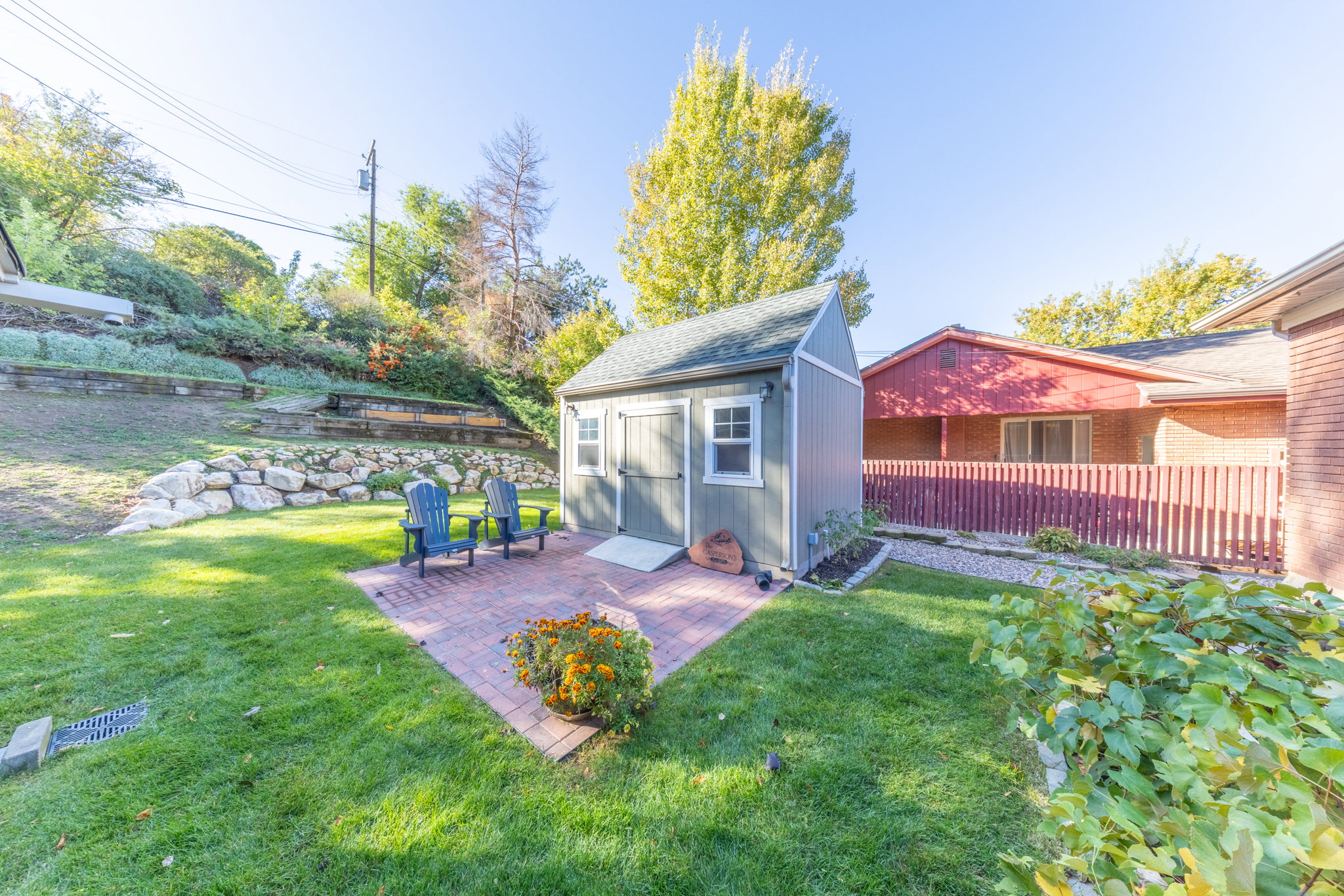 Backyard featuring a shed with a gable roof, a brick paver patio, a terraced landscape with rock retaining walls, and mature trees