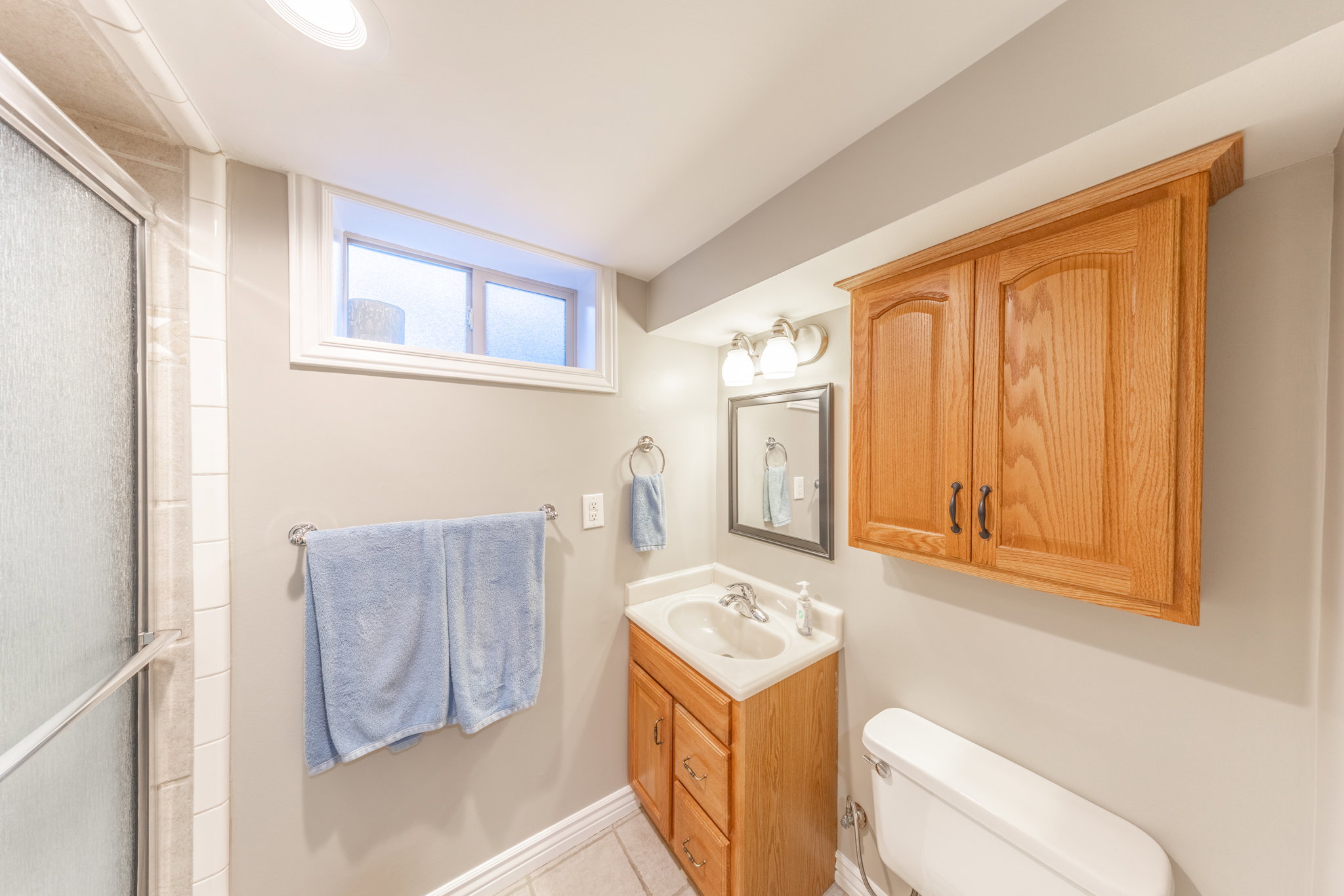 Bathroom featuring a shower with glass enclosure, wood-finish vanity with a white basin, framed mirror, and overhead wood-finish cabinetry