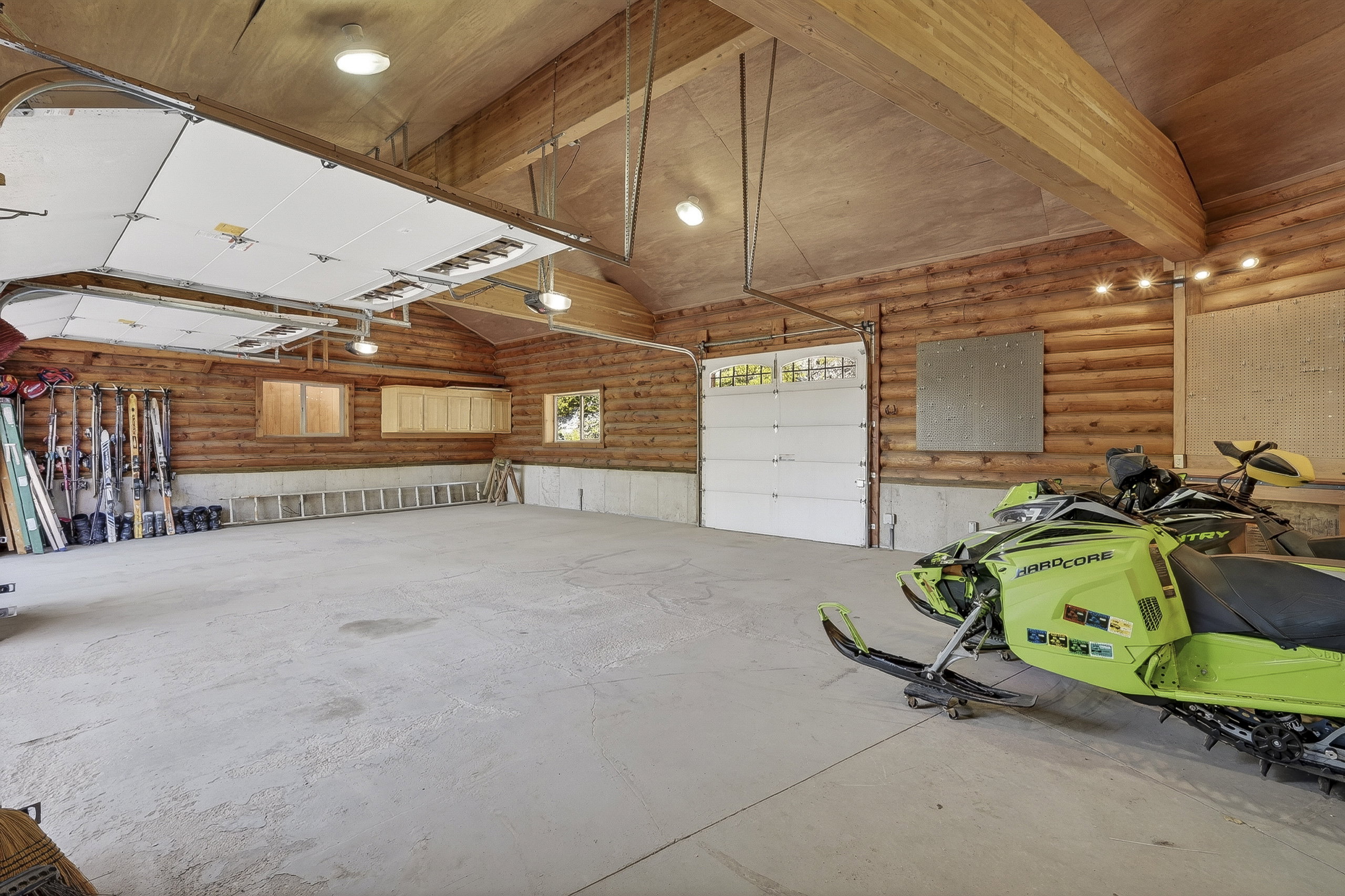Spacious garage featuring log wall construction, exposed wood beam ceiling, concrete flooring, dual overhead garage doors, and built-in cabinetry