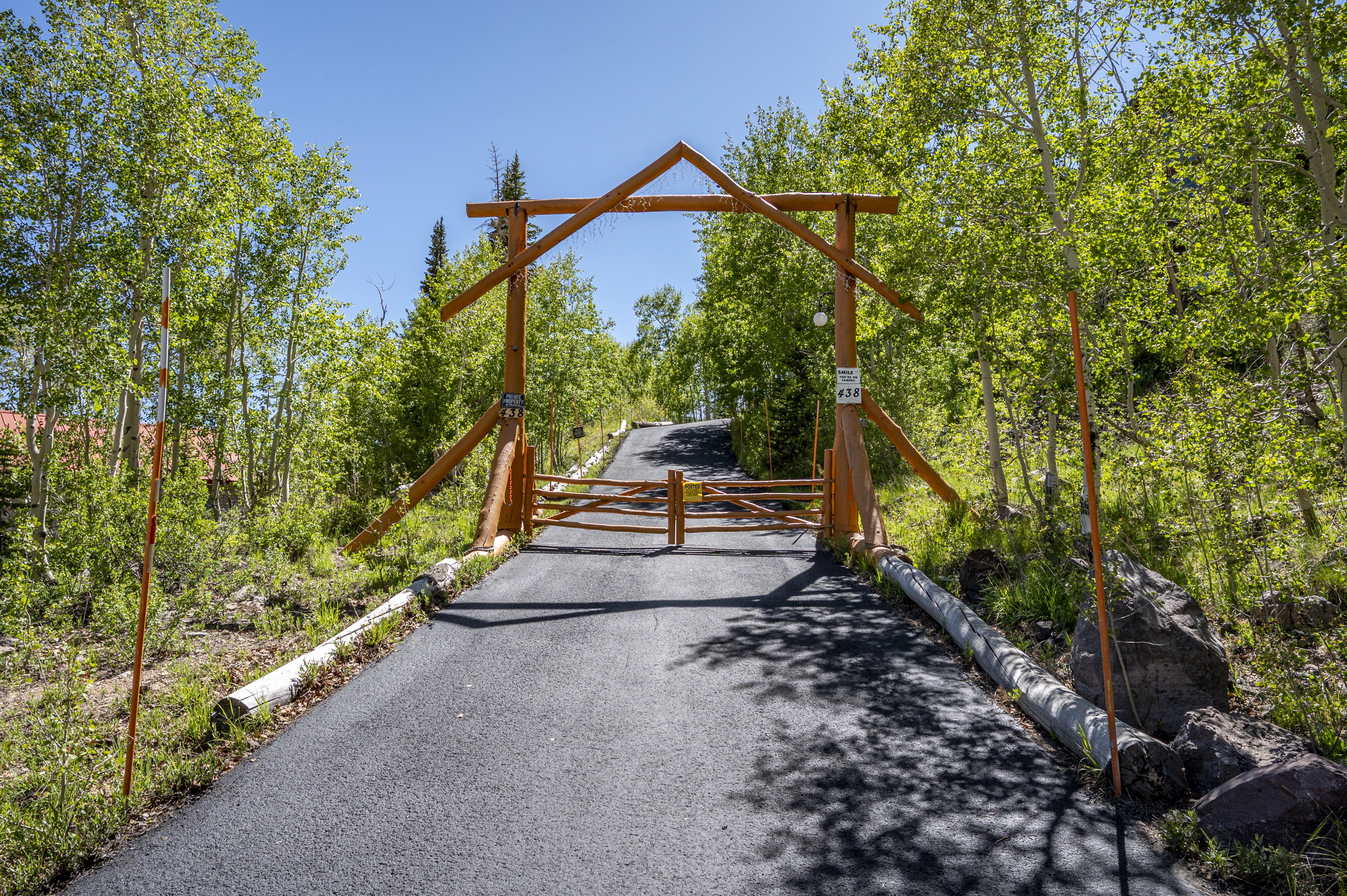 Paved driveway entrance with a rustic wooden gate and overhead archway, framed by mature trees