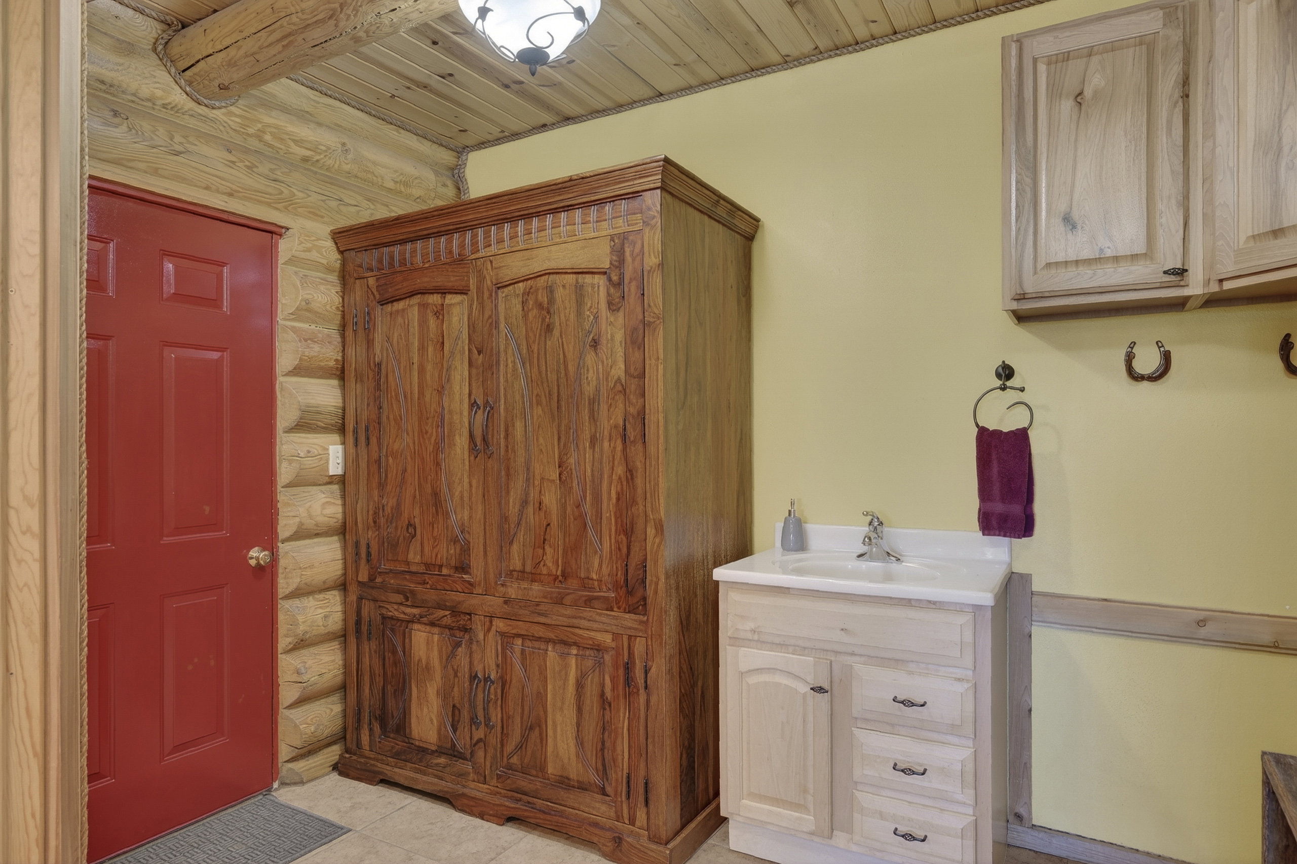 Log cabin interior featuring a red paneled door, a large wood-finish armoire, a light wood-finish vanity with a white countertop sink, and overhead cabinetry