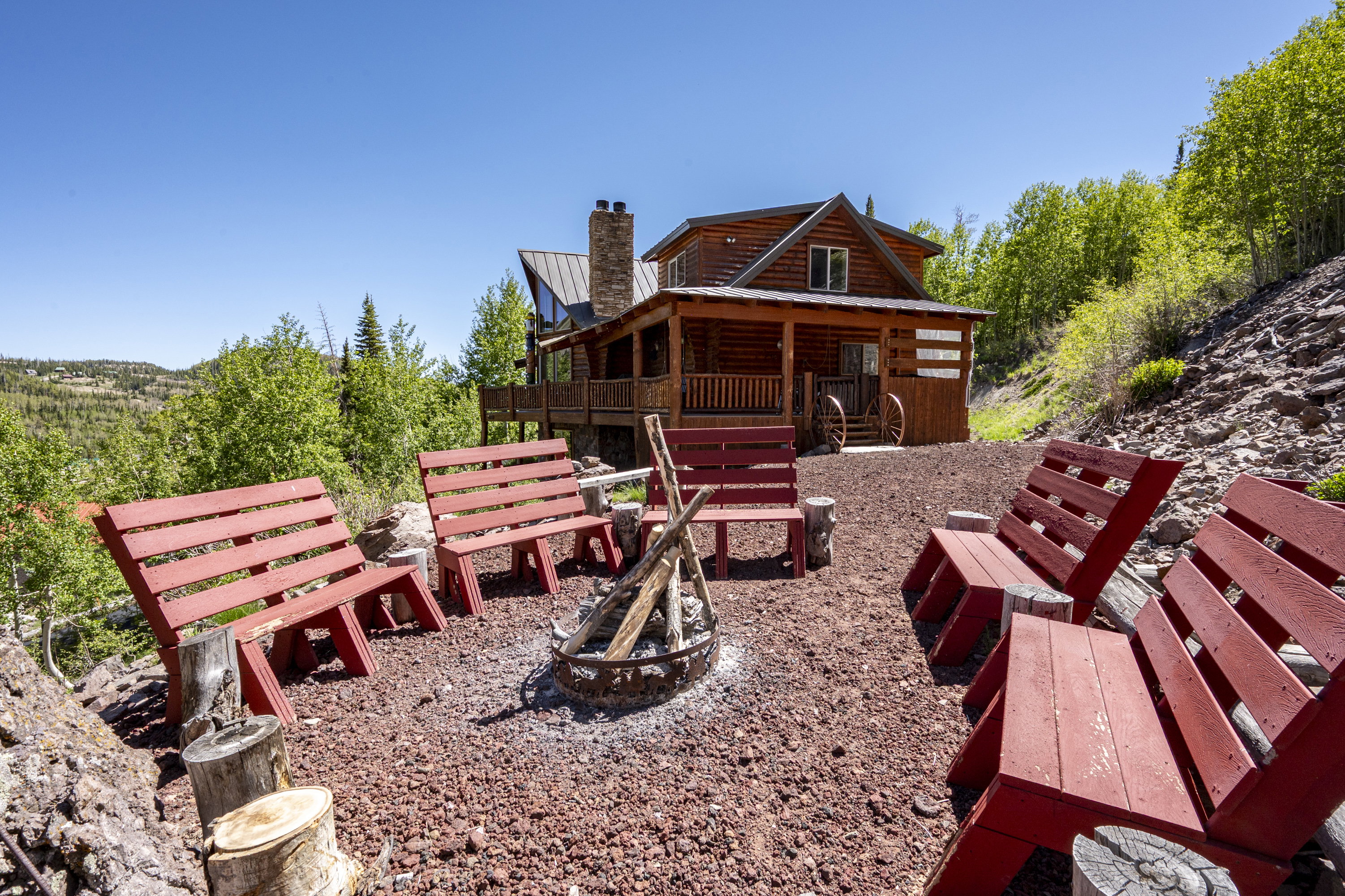 Outdoor fire pit area with red benches and a gravel base, set against a backdrop of mature trees