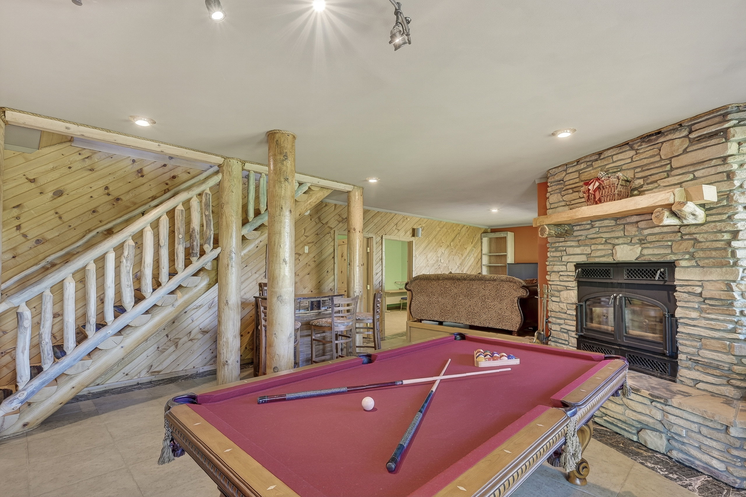 Spacious entertainment area featuring a stone-clad fireplace with a wood mantel, wood-paneled walls, a rustic log staircase, and recessed lighting