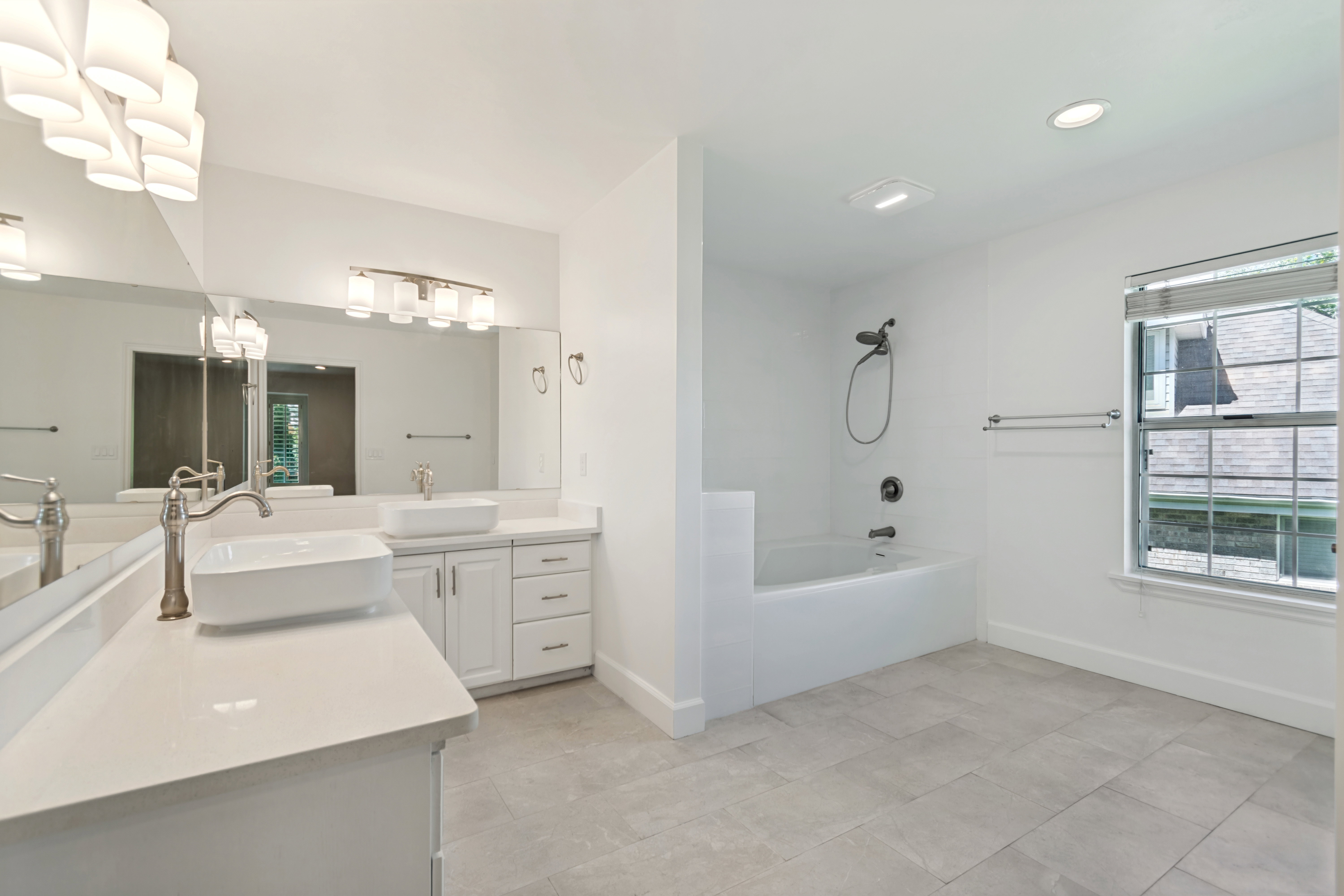 Bathroom featuring dual vessel sinks, white cabinetry, a built-in bathtub with shower, light-toned tile flooring, and a window with blinds