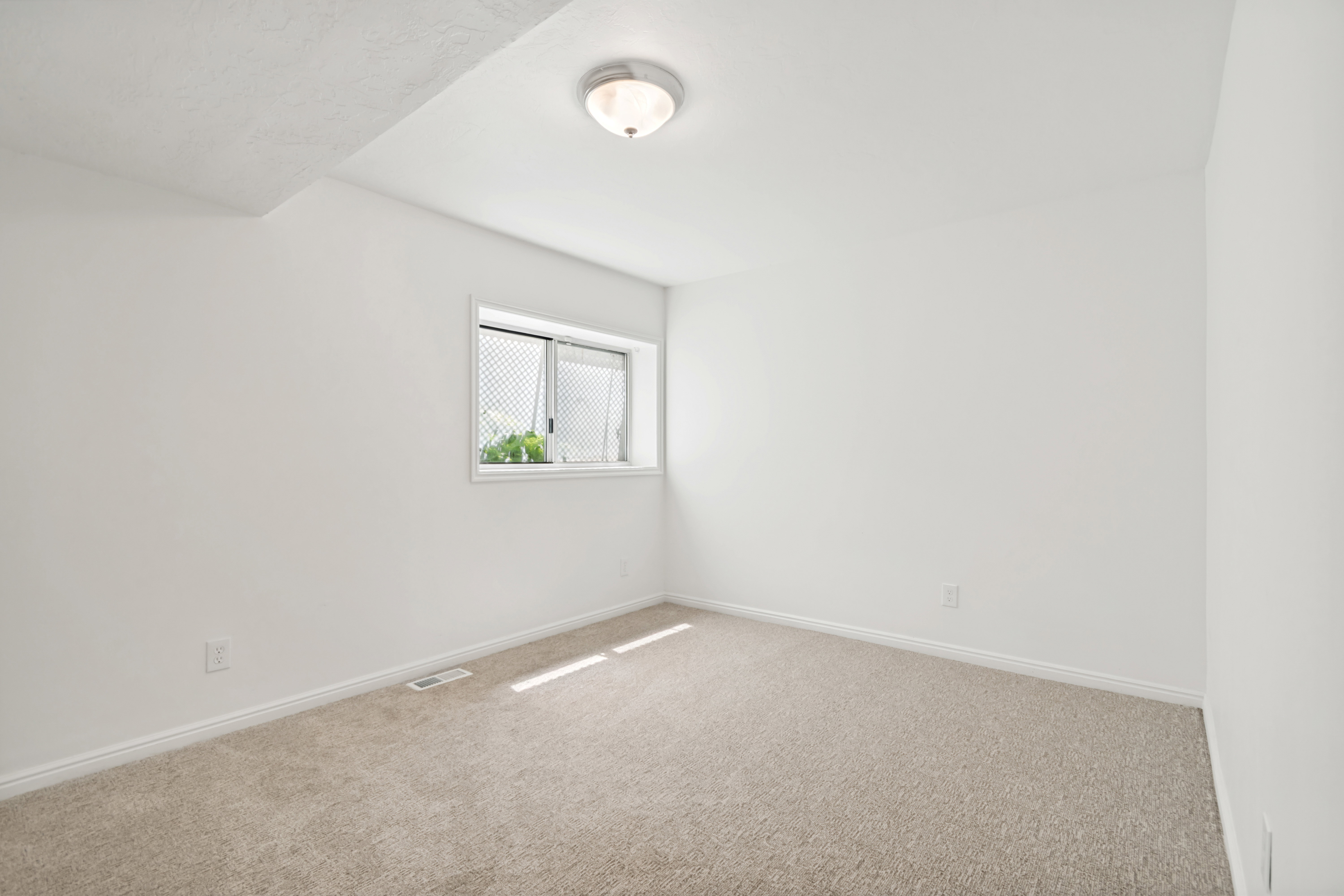 Bright room featuring a window with security grating, neutral-toned carpet, and white baseboards