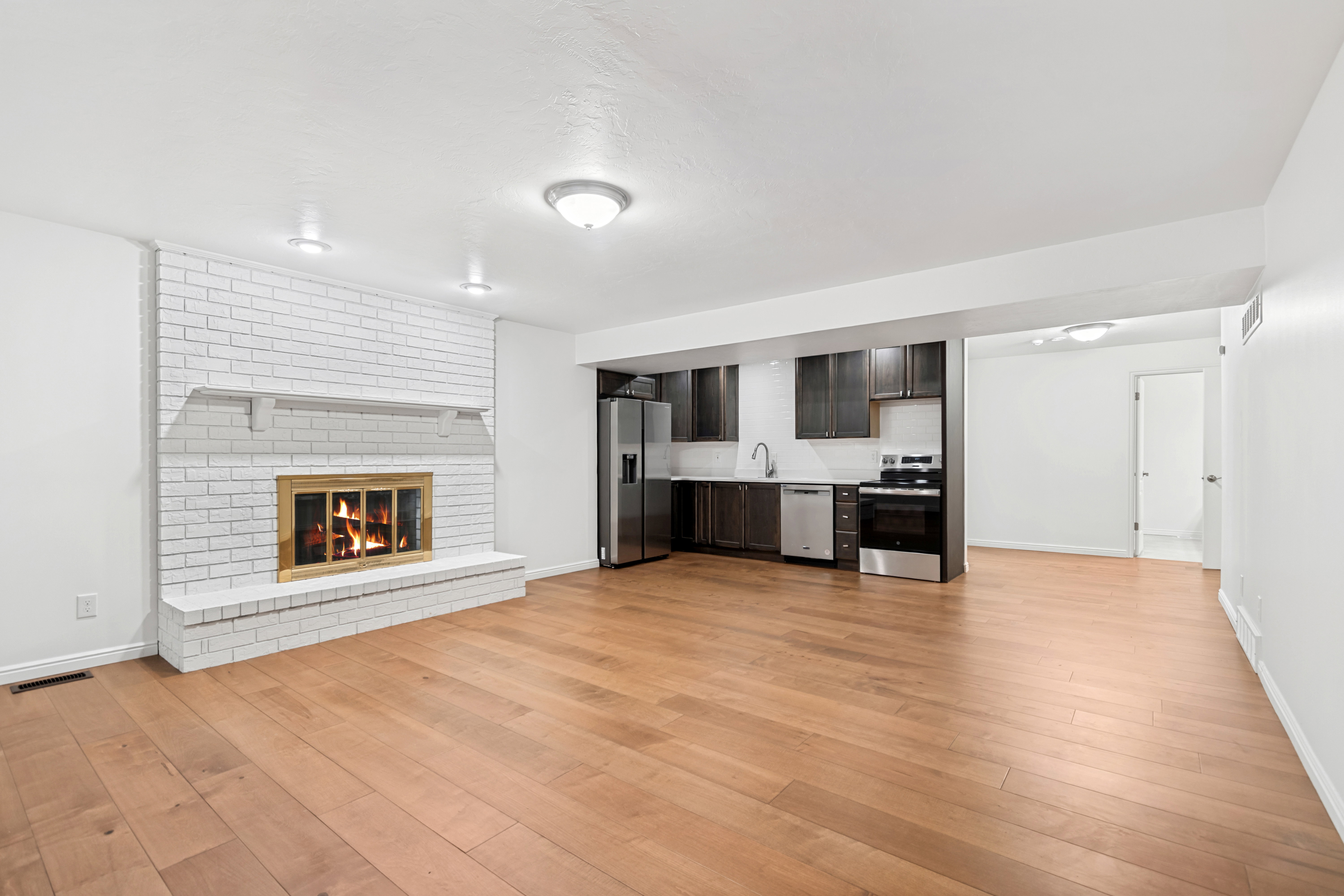 Spacious room featuring wide plank wood-finish flooring, a white painted brick fireplace with a brass-trimmed firebox, recessed lighting, and a compact kitchen area