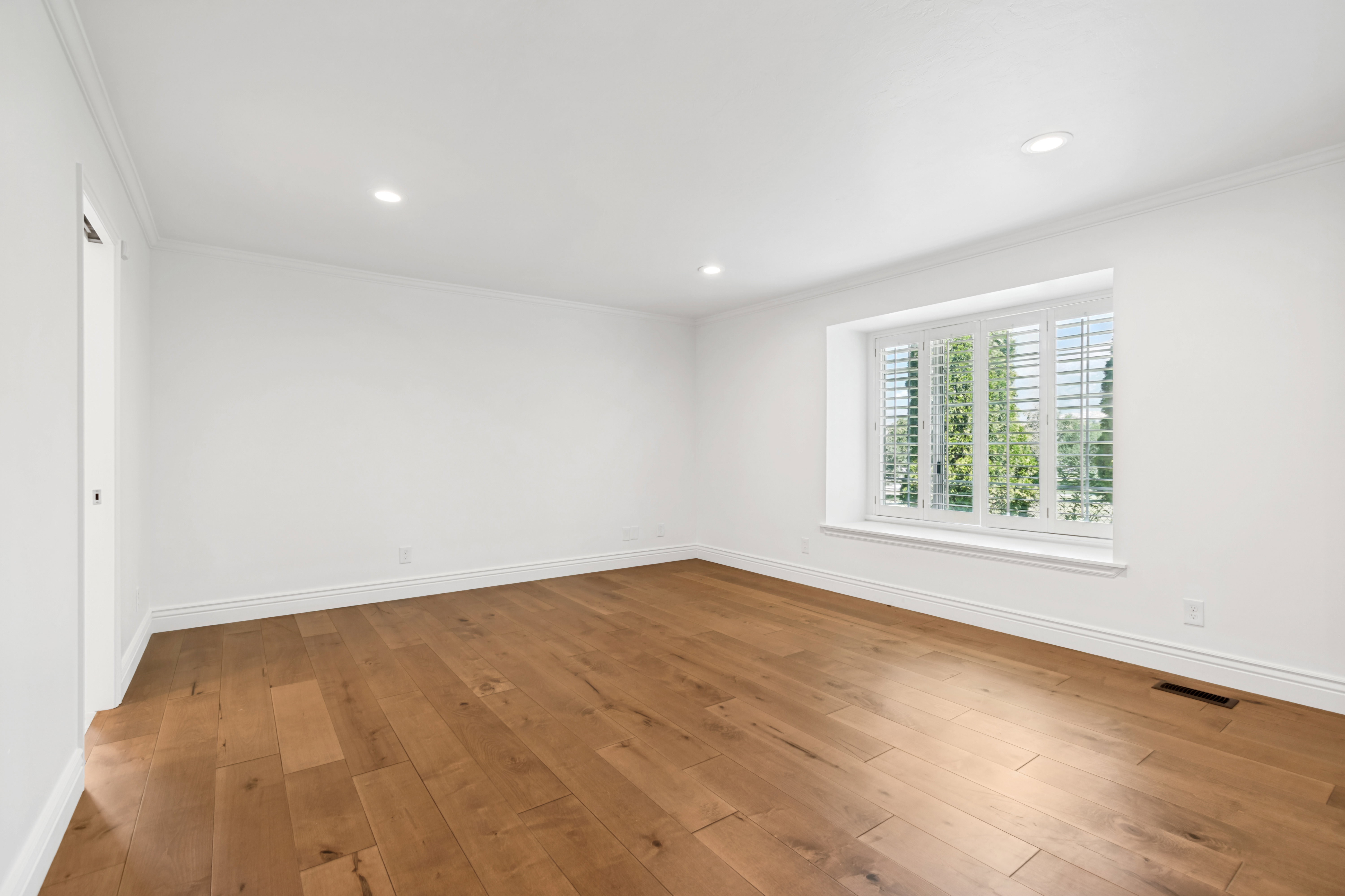 Spacious room featuring wood-finish flooring, recessed lighting, crown molding, and a window with white plantation shutters