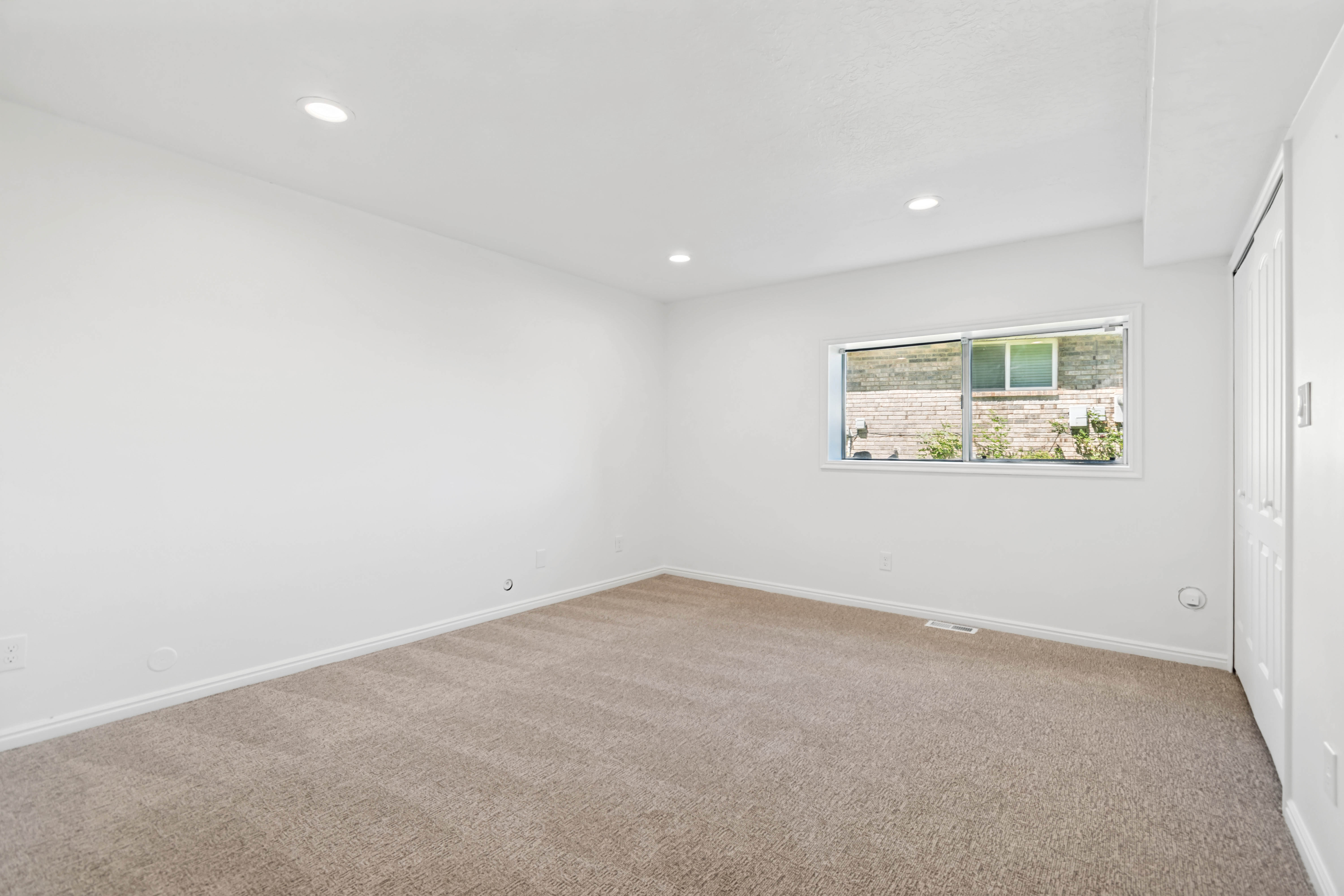 Spacious room featuring neutral carpeting, white walls, recessed ceiling lighting, a window with a white frame, and a bi-fold closet door