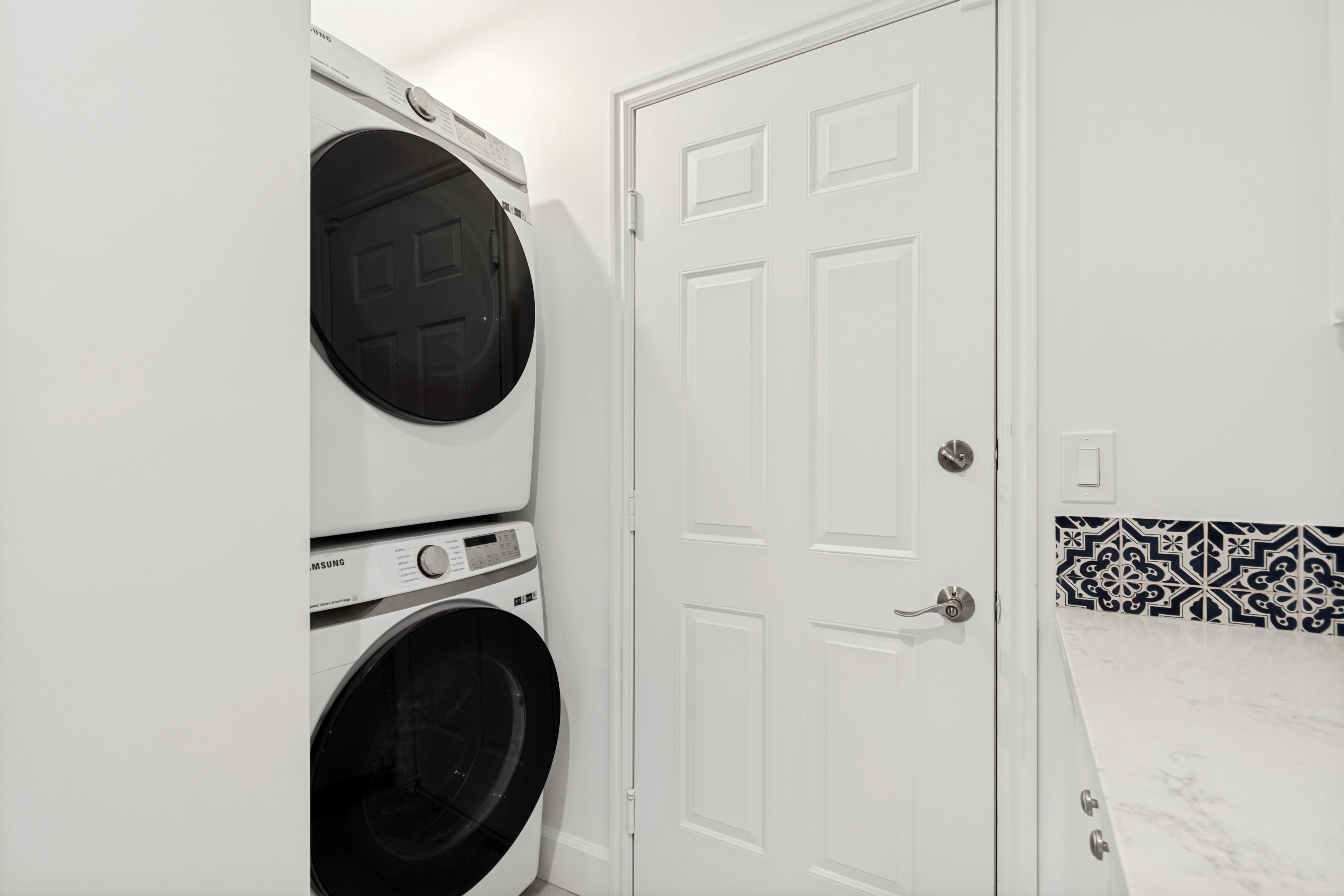 Laundry area featuring a stacked washer and dryer, patterned accent tile, a solid white door with brushed nickel hardware, and a white countertop