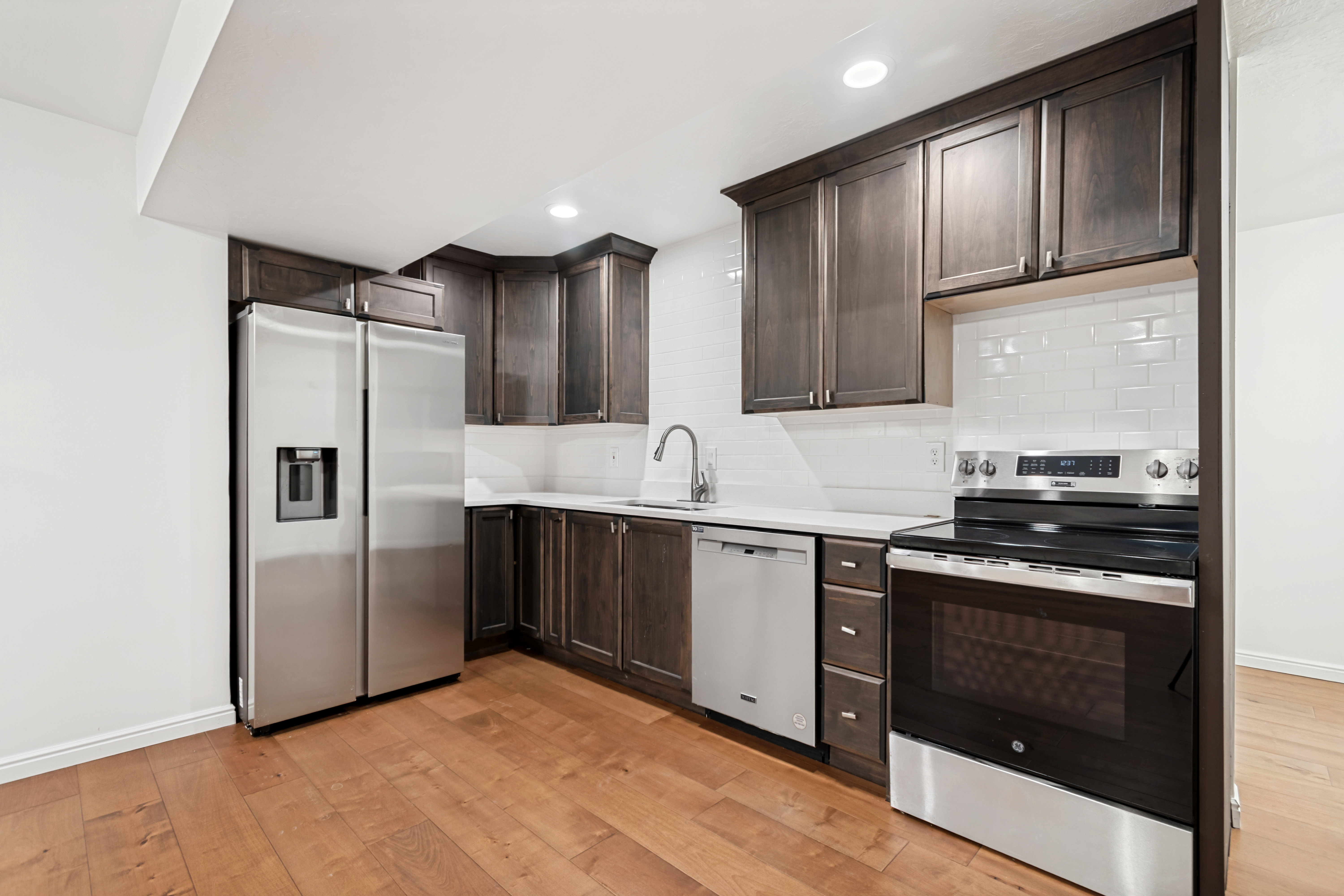 Modern kitchen featuring dark wood cabinetry, stainless steel appliances, white subway tile backsplash, light countertops, and wood-finish flooring