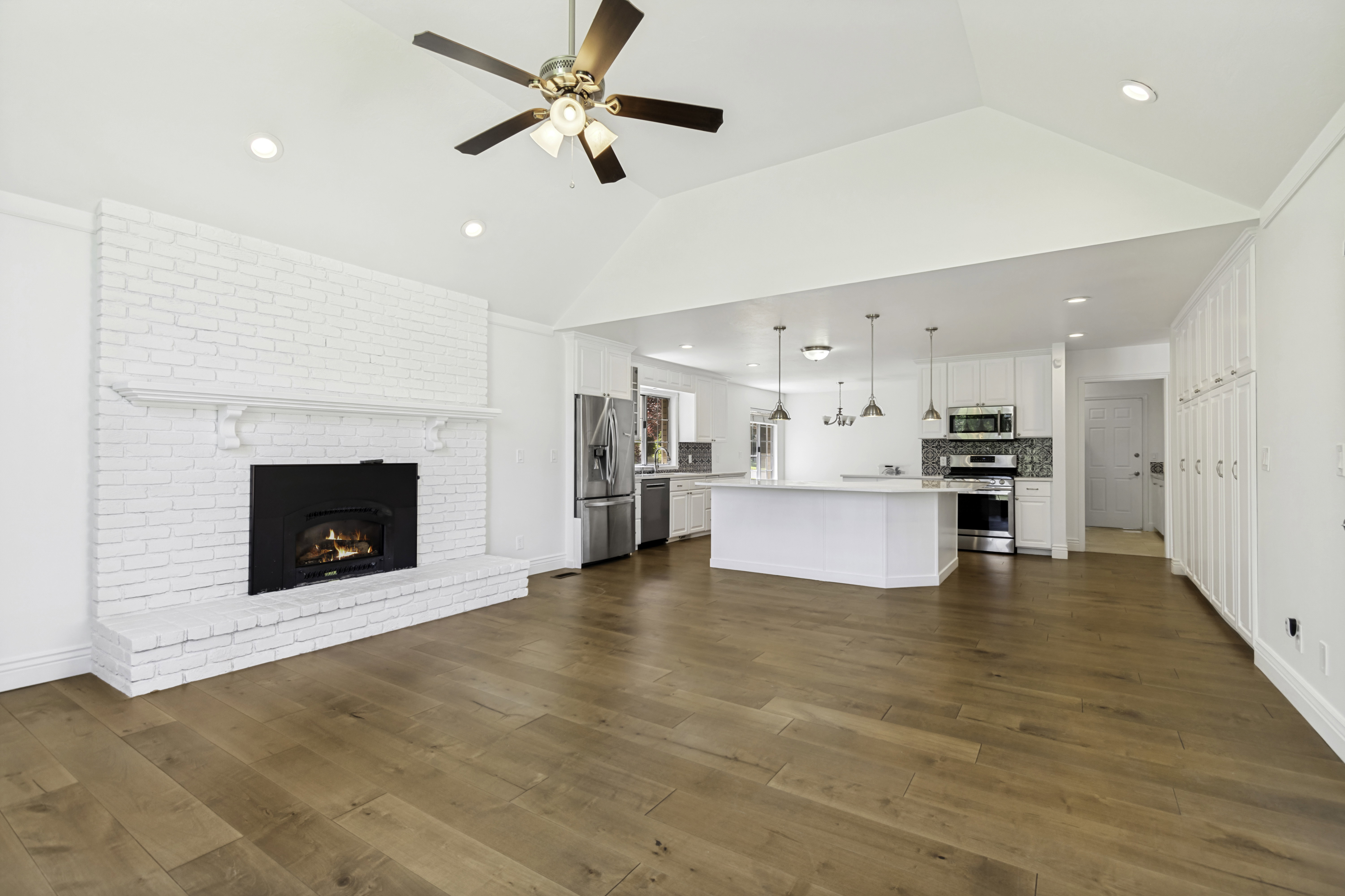 Expansive living area featuring hardwood flooring, a vaulted ceiling, and a white brick fireplace with a mantel