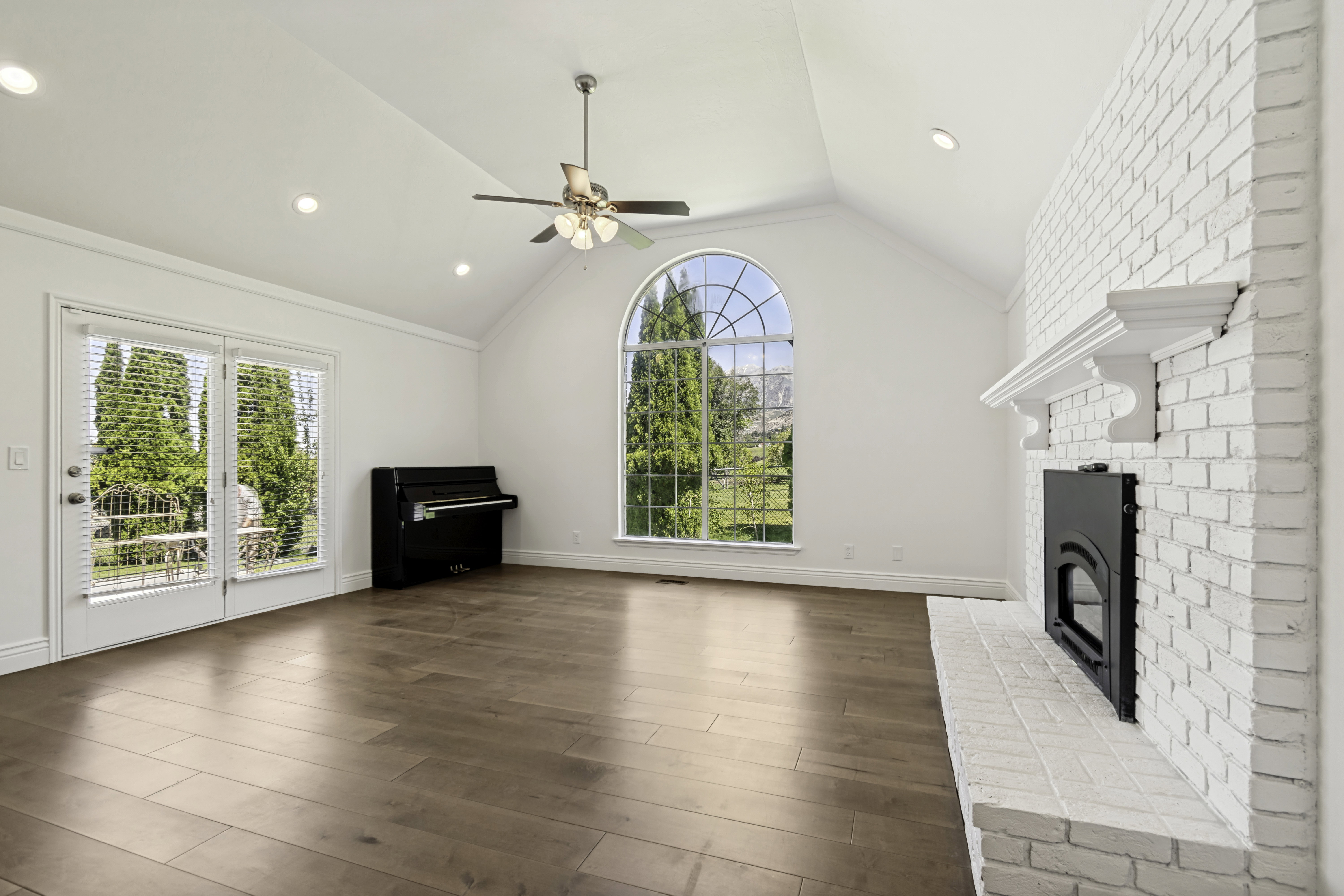Spacious living area featuring vaulted ceilings, wood-finish flooring, a white brick fireplace with a mantel, and an arched picture window