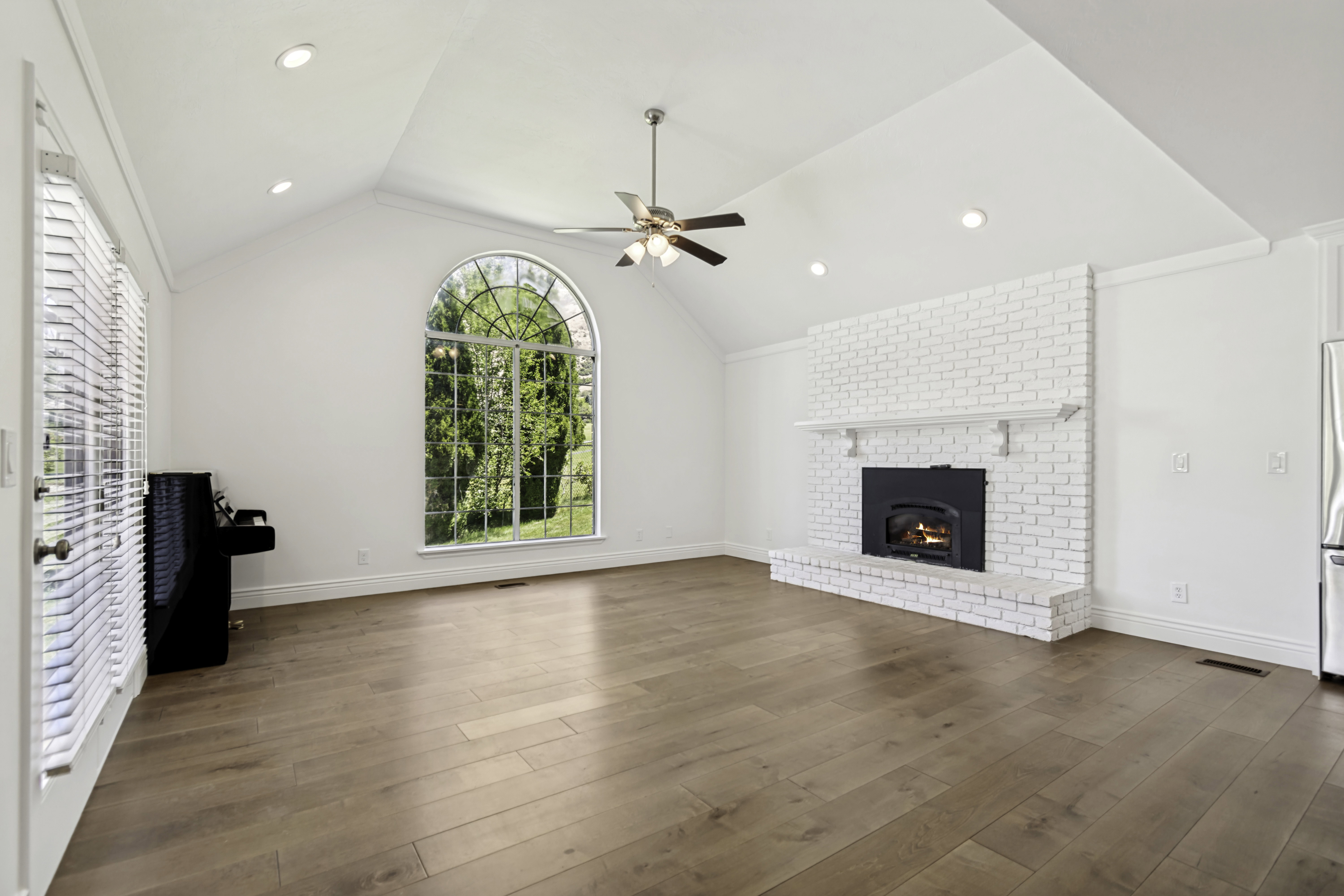 Vaulted ceiling great room featuring wood-finish flooring, a white-painted brick fireplace with mantel, and a large arched window