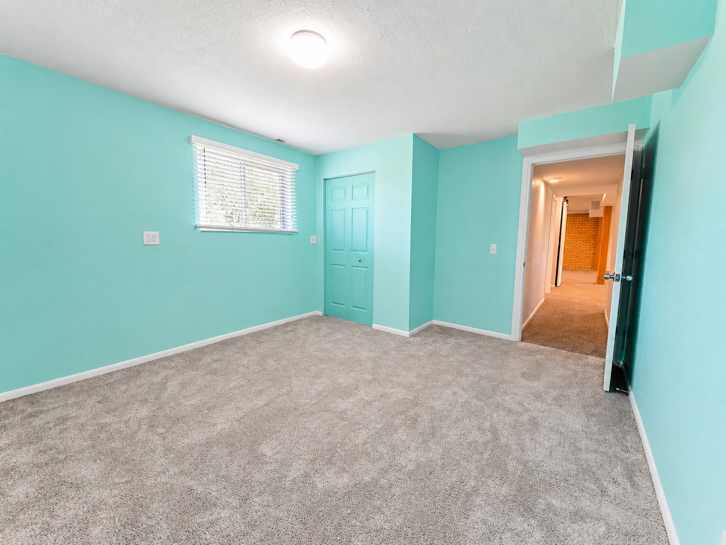 Spacious room featuring light blue walls, plush gray carpeting, a window with horizontal blinds, and a bi-fold closet door