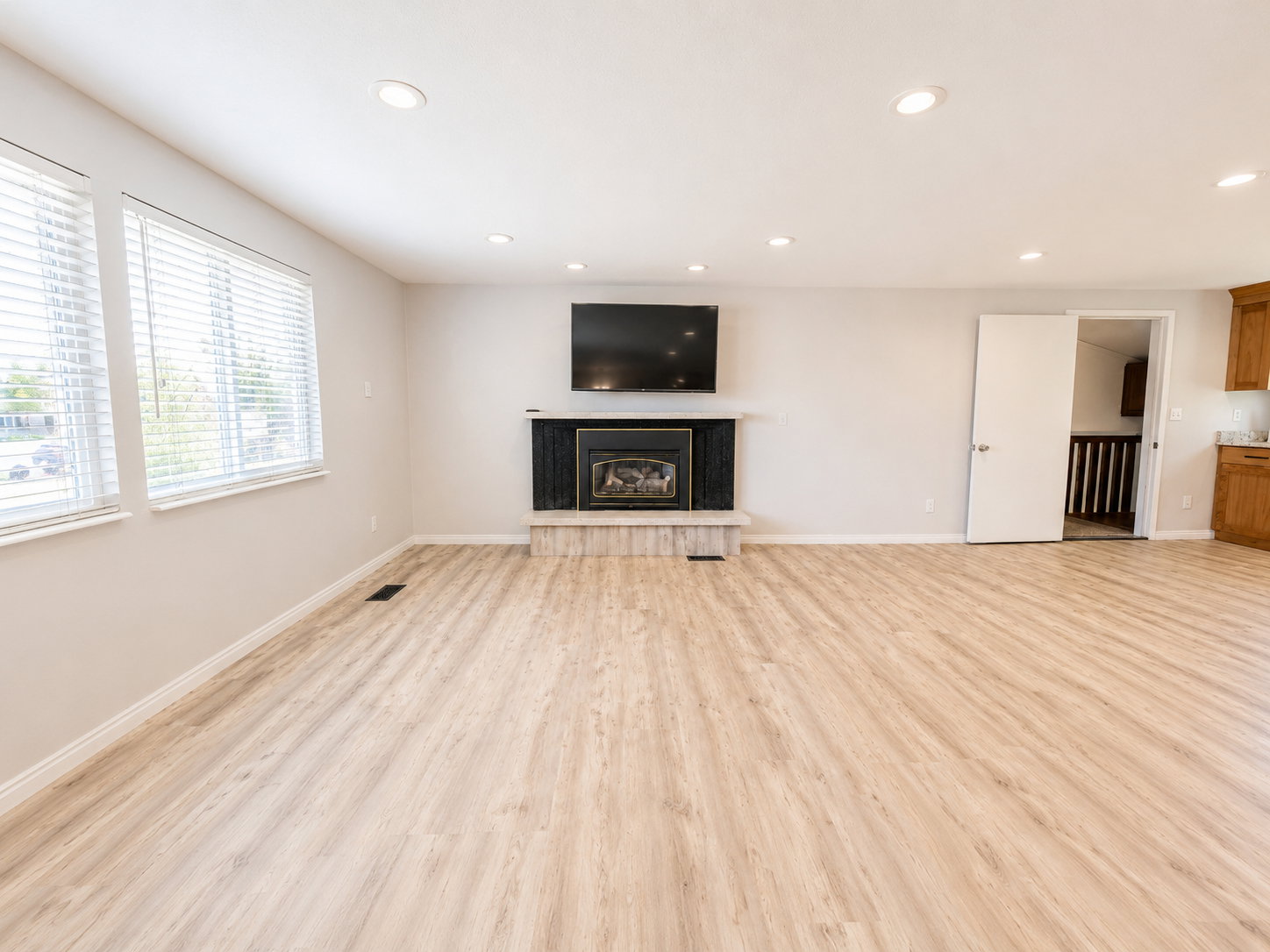 Spacious room featuring wood-finish flooring, recessed ceiling lighting, a fireplace with a black surround and stone hearth, and multiple windows with blinds