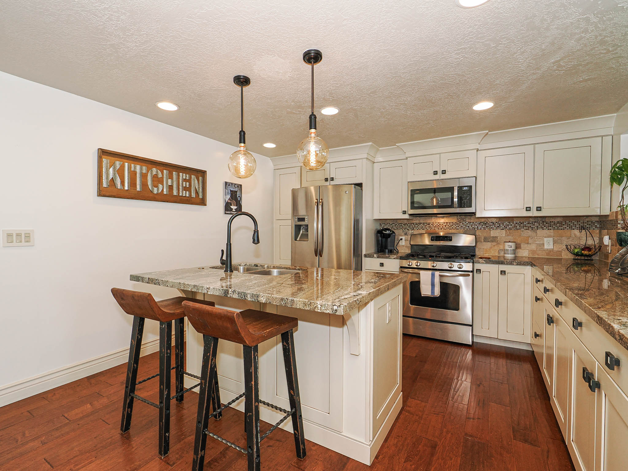 Kitchen featuring a center island with a built-in sink, pendant lighting, stainless steel appliances, white cabinetry with dark hardware, and wood-finish flooring