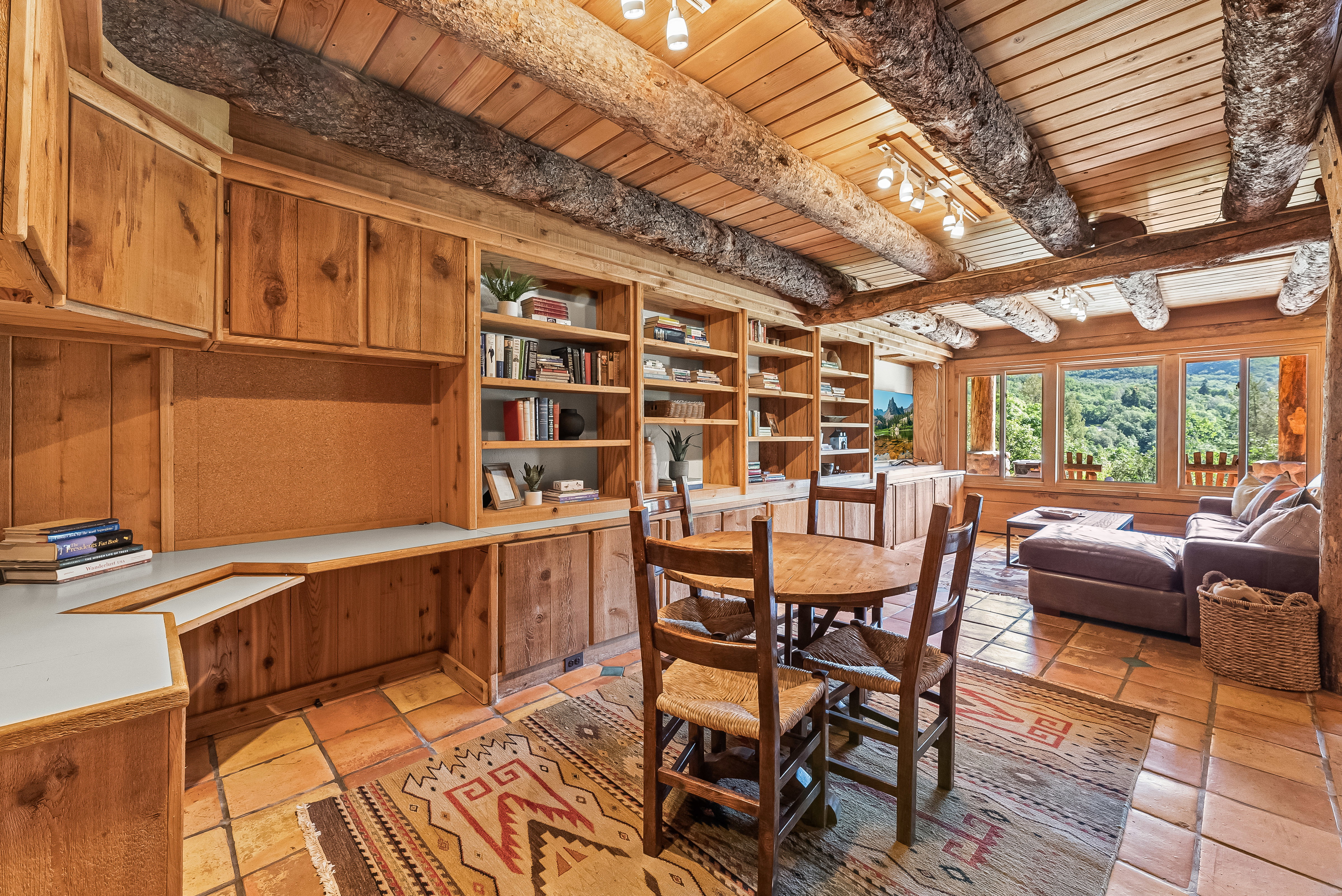 Expansive room featuring natural wood walls and ceiling, exposed log beams, built-in shelving, a dedicated desk area with upper cabinetry, and large windows offering abundant natural light