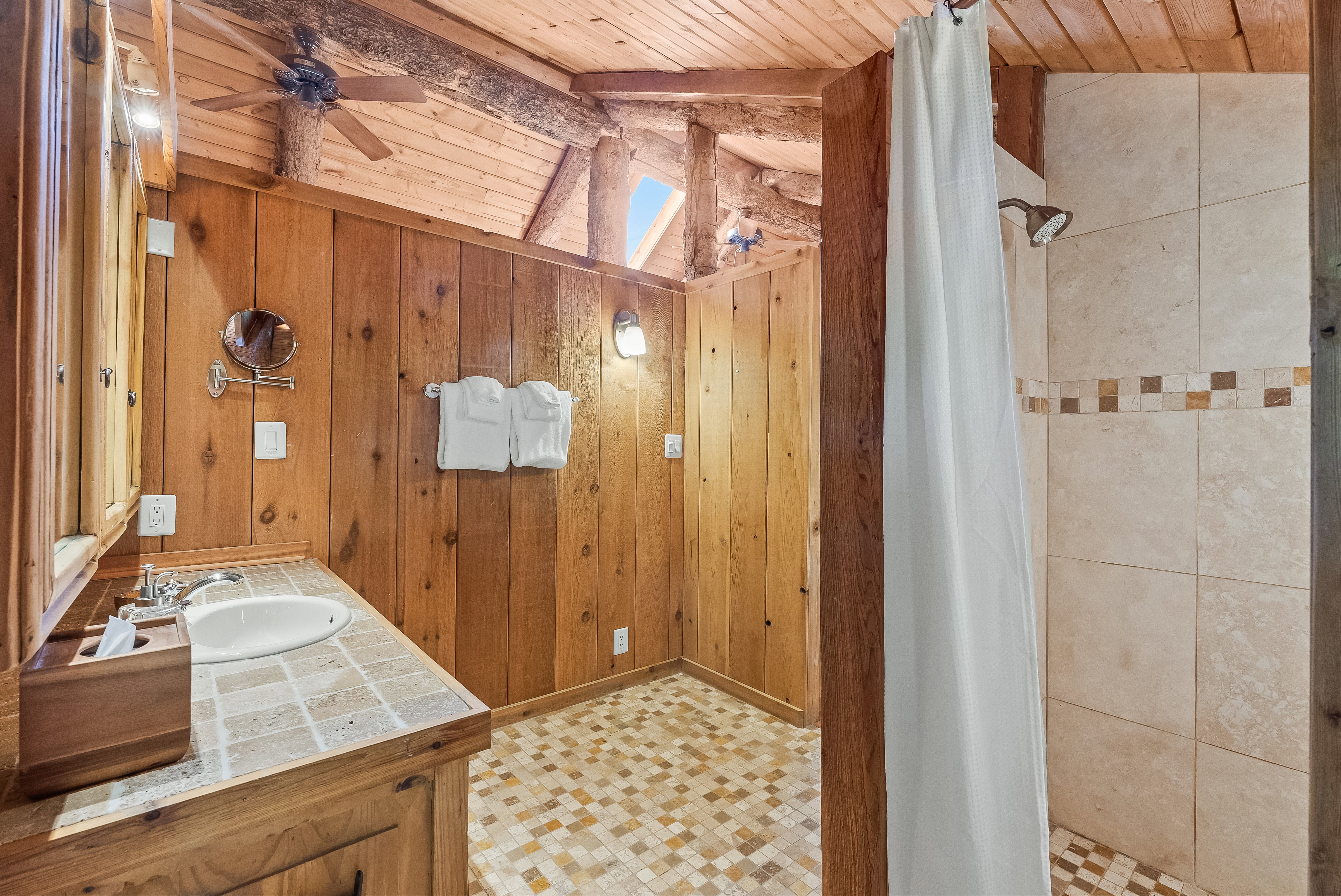 Rustic bathroom featuring wood-paneled walls and ceiling, a wood vanity with a tile countertop and integrated sink, and a tiled shower with an accent band