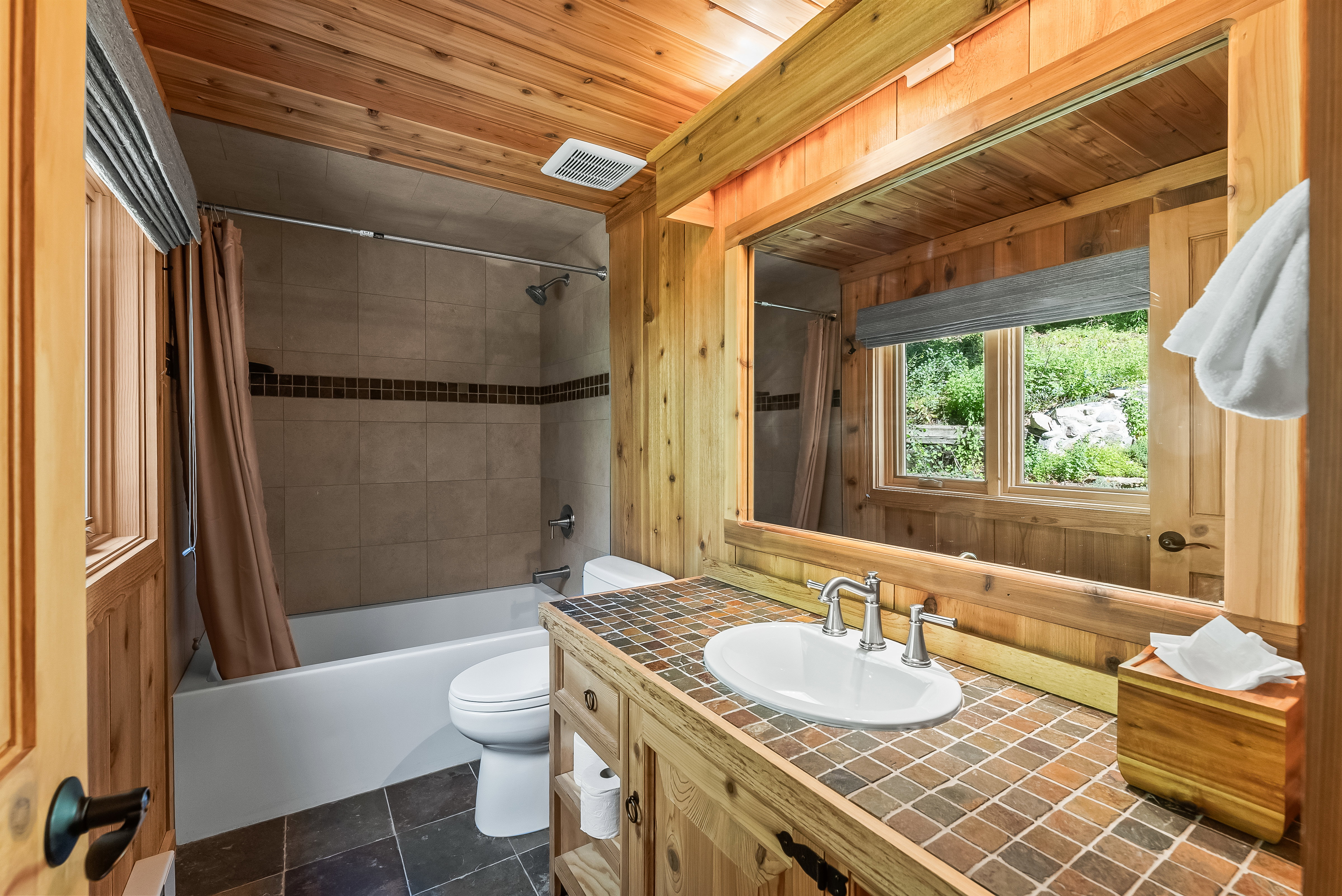 Bathroom featuring extensive wood paneling, a single vanity with a tile countertop, an oval undermount sink, and a framed mirror
