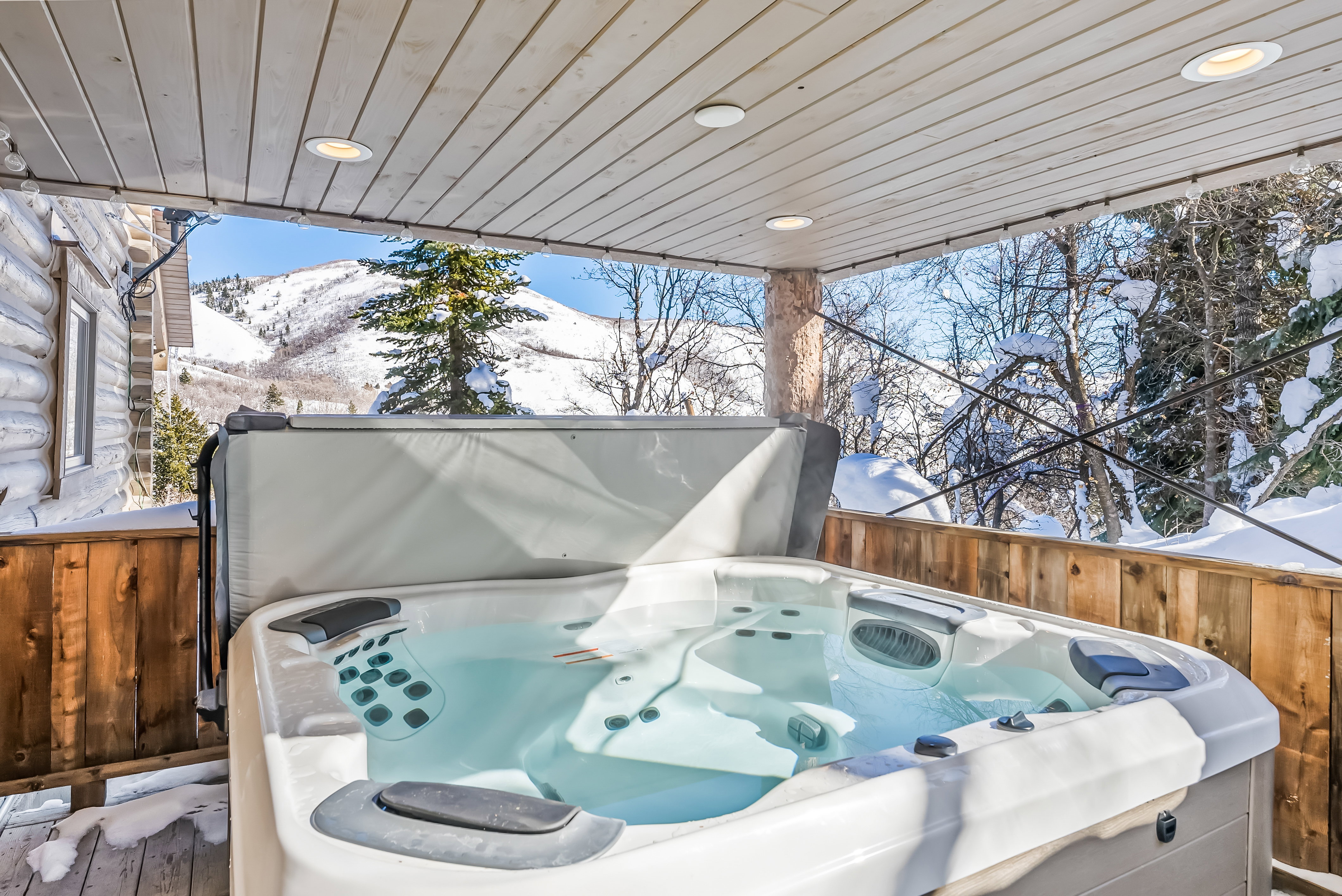 Covered outdoor hot tub with mountain scenery, log cabin exterior, wood-finish railing, and recessed ceiling lighting
