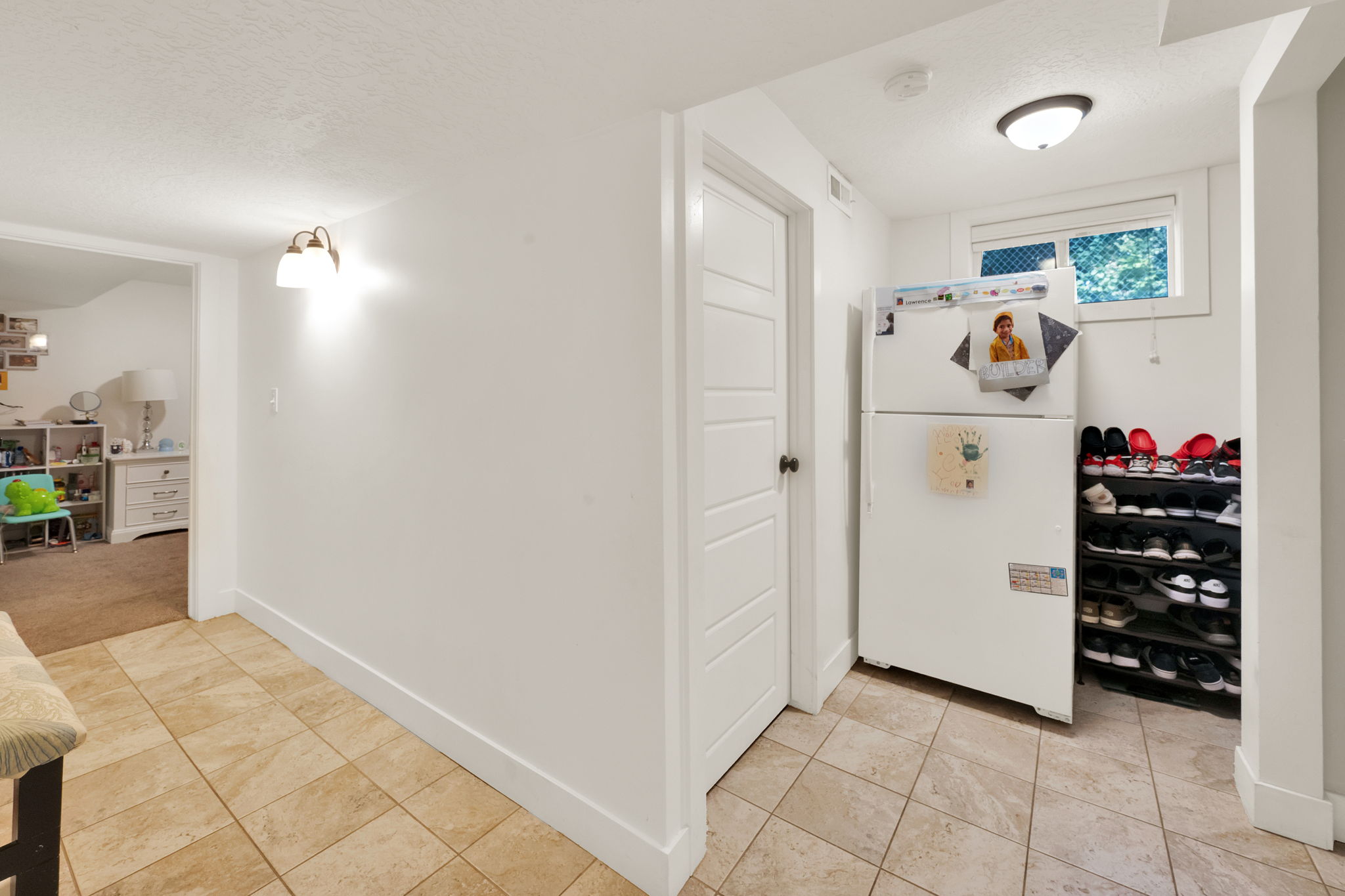 Tiled flooring extending into a utility area with a window, a white door, and a wall-mounted light fixture