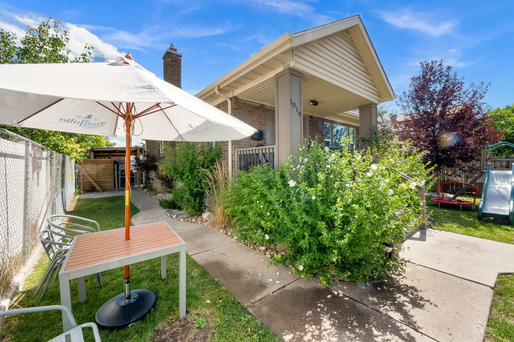 Manicured backyard featuring a concrete patio, established plantings, mature trees, and a chain-link fence