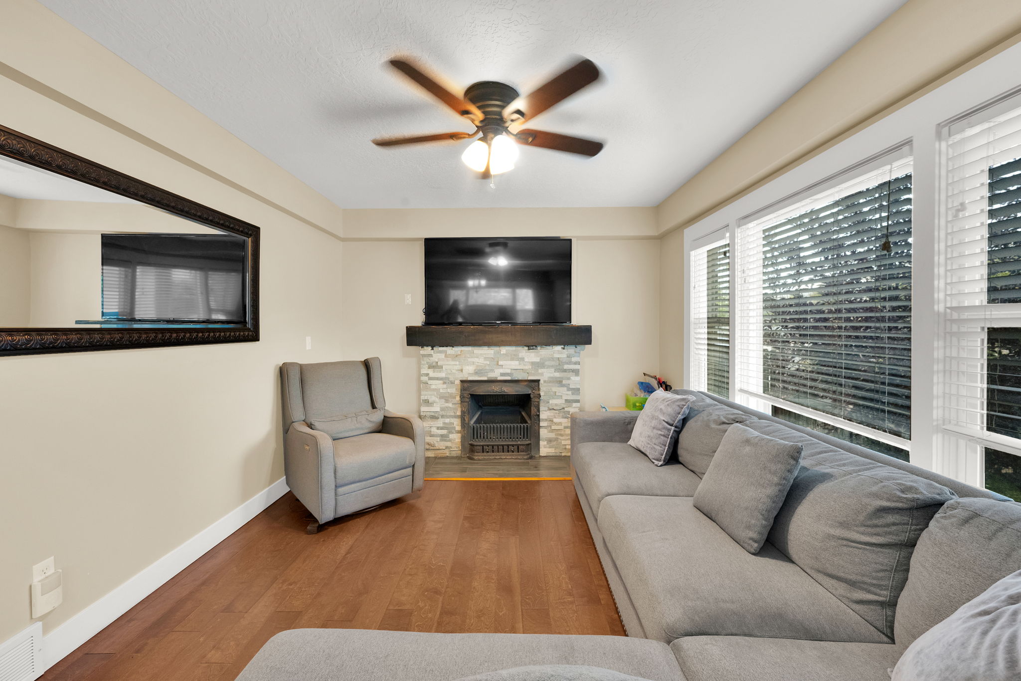 Living area featuring wood-finish flooring, a stacked stone fireplace with wood mantel, a ceiling fan with integrated lighting, and multiple large windows