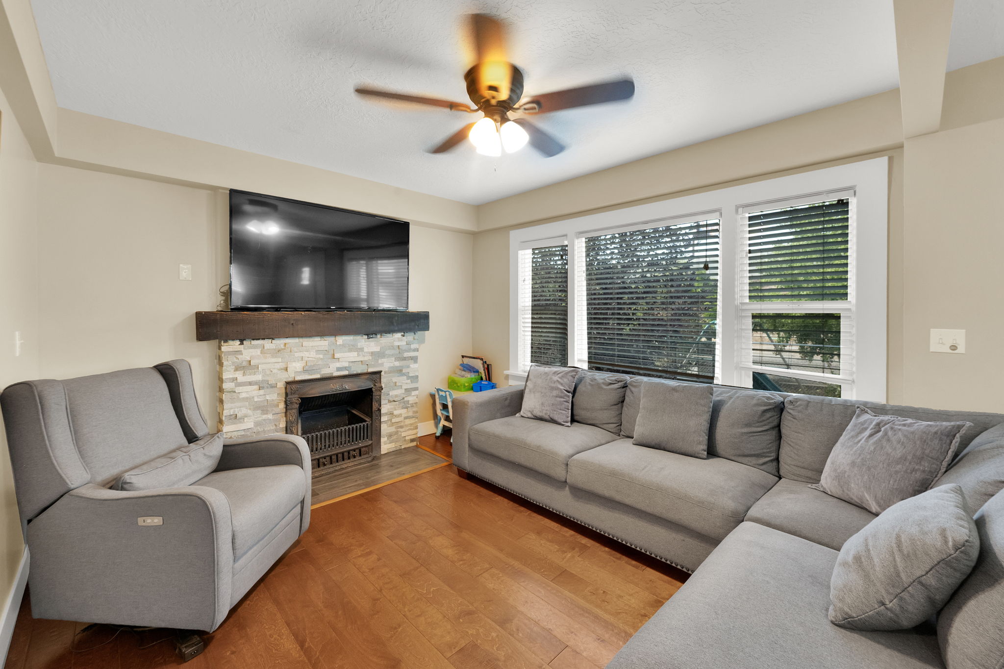 Living area featuring wood-finish flooring, a stacked stone fireplace with a substantial wood mantel, multiple large windows with blinds, a ceiling fan with integrated lighting, and neutral wall tones