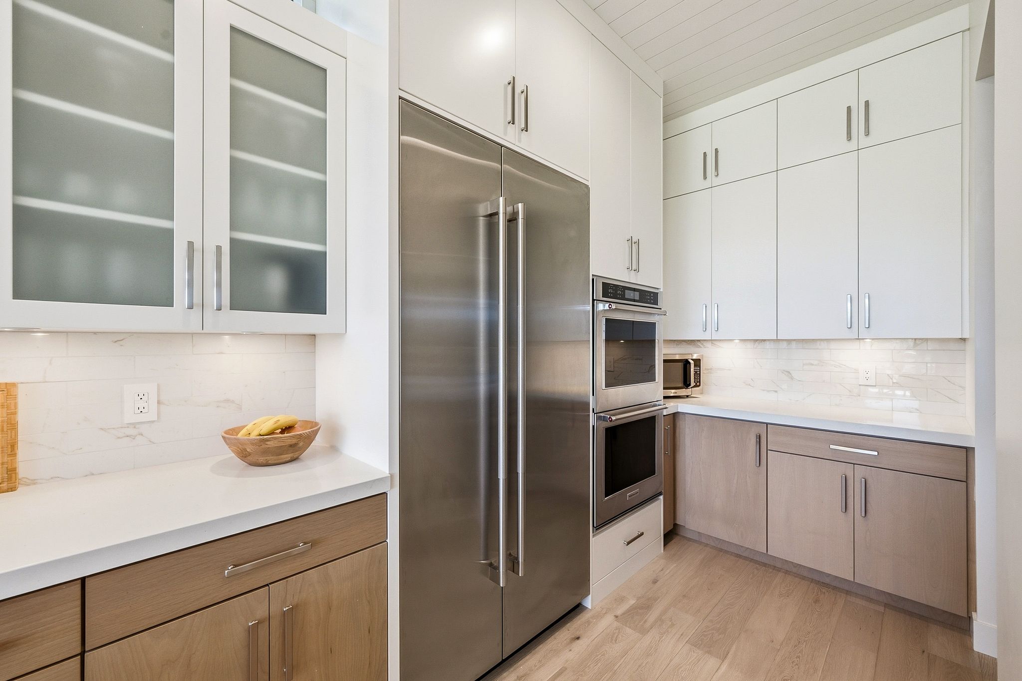 Contemporary kitchen featuring a full-size stainless steel refrigerator, double wall ovens, white upper cabinetry with some frosted glass fronts, and light wood-finish lower cabinetry, all complemented by white countertops and a tile backsplash