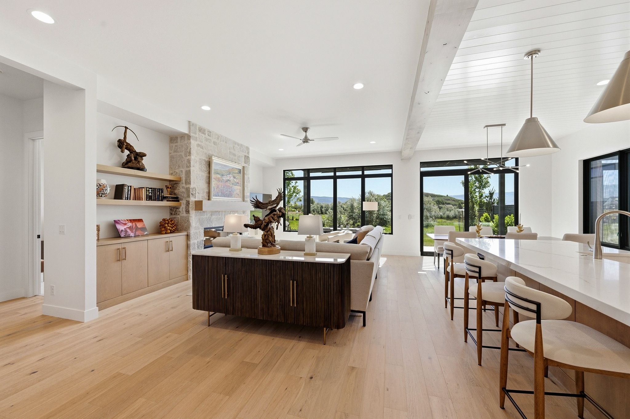 Open-concept interior featuring light wood-finish flooring, a stone accent wall with a fireplace, built-in shelving, and extensive black-framed windows