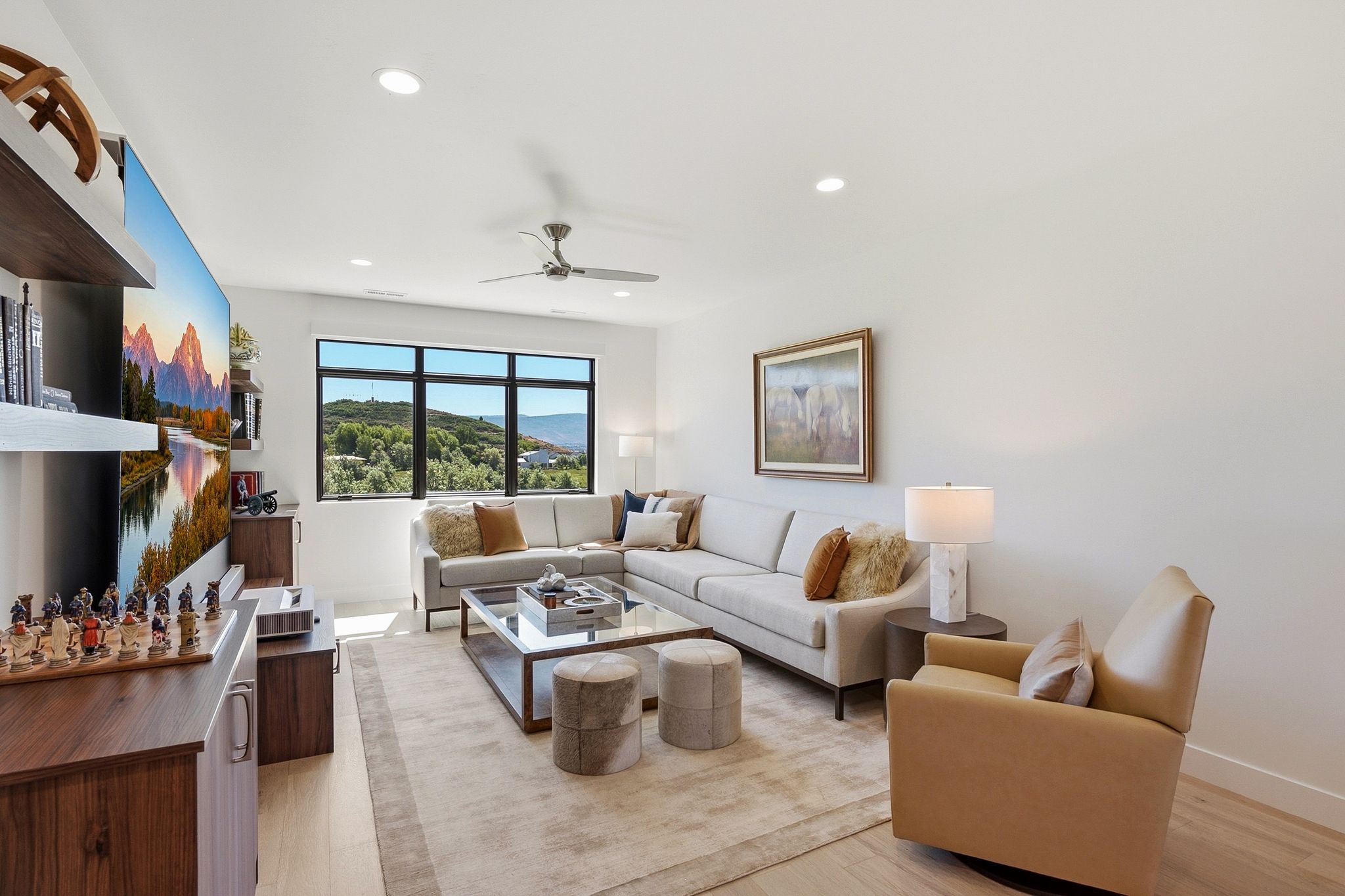 Spacious living area featuring large windows showcasing surrounding scenery, light wood-finish flooring, recessed lighting, and a ceiling fan