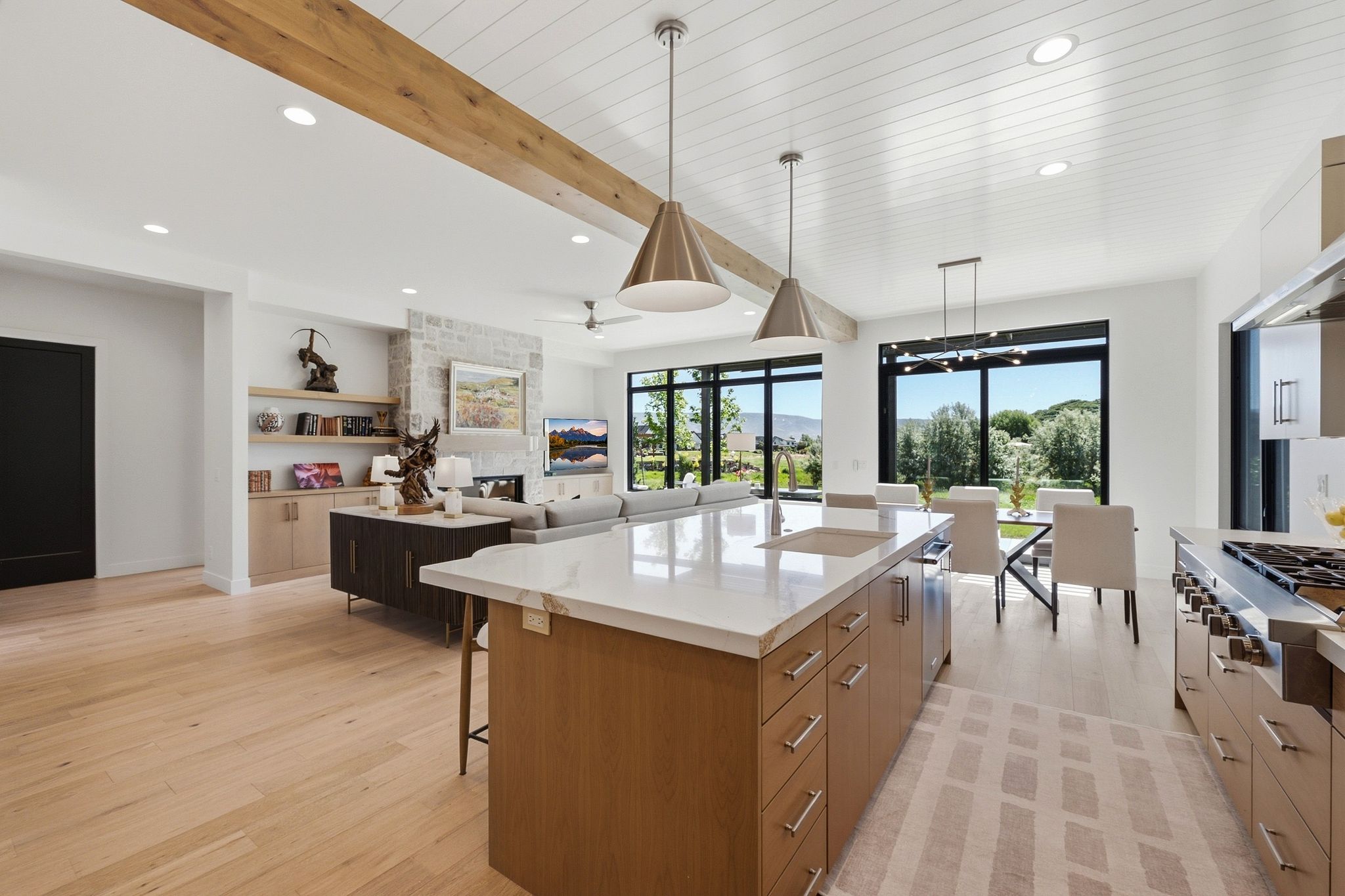 Open-concept kitchen and living area featuring light wood-finish flooring, a kitchen island with a built-in sink and light-toned countertop, a range with stainless steel hood, exposed wood beam ceiling accents, and large windows