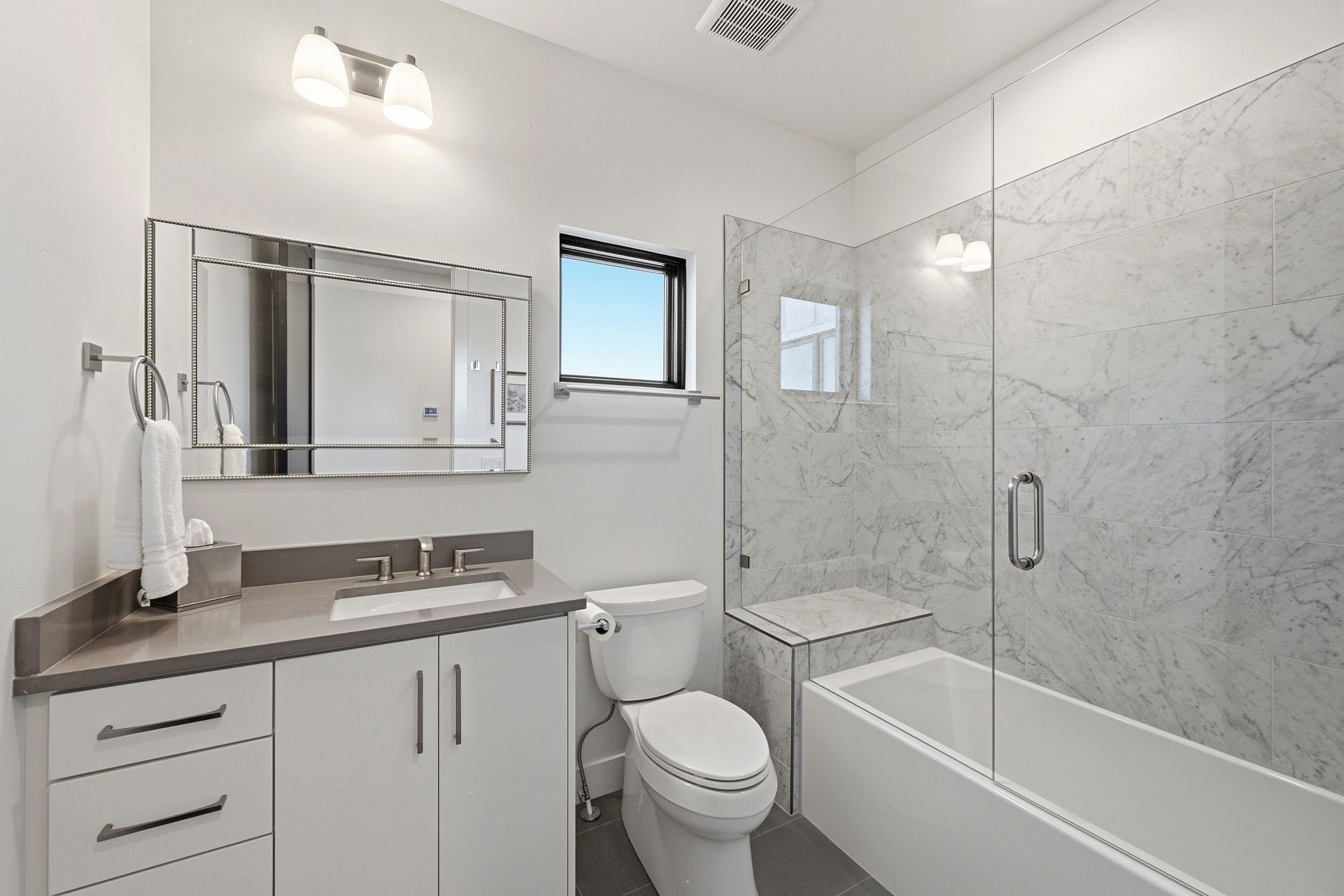 Full bathroom featuring a rectangular vanity mirror, a single vanity with a gray countertop and undermount sink, and a combination tub/shower with a glass enclosure and marble-look tile surround