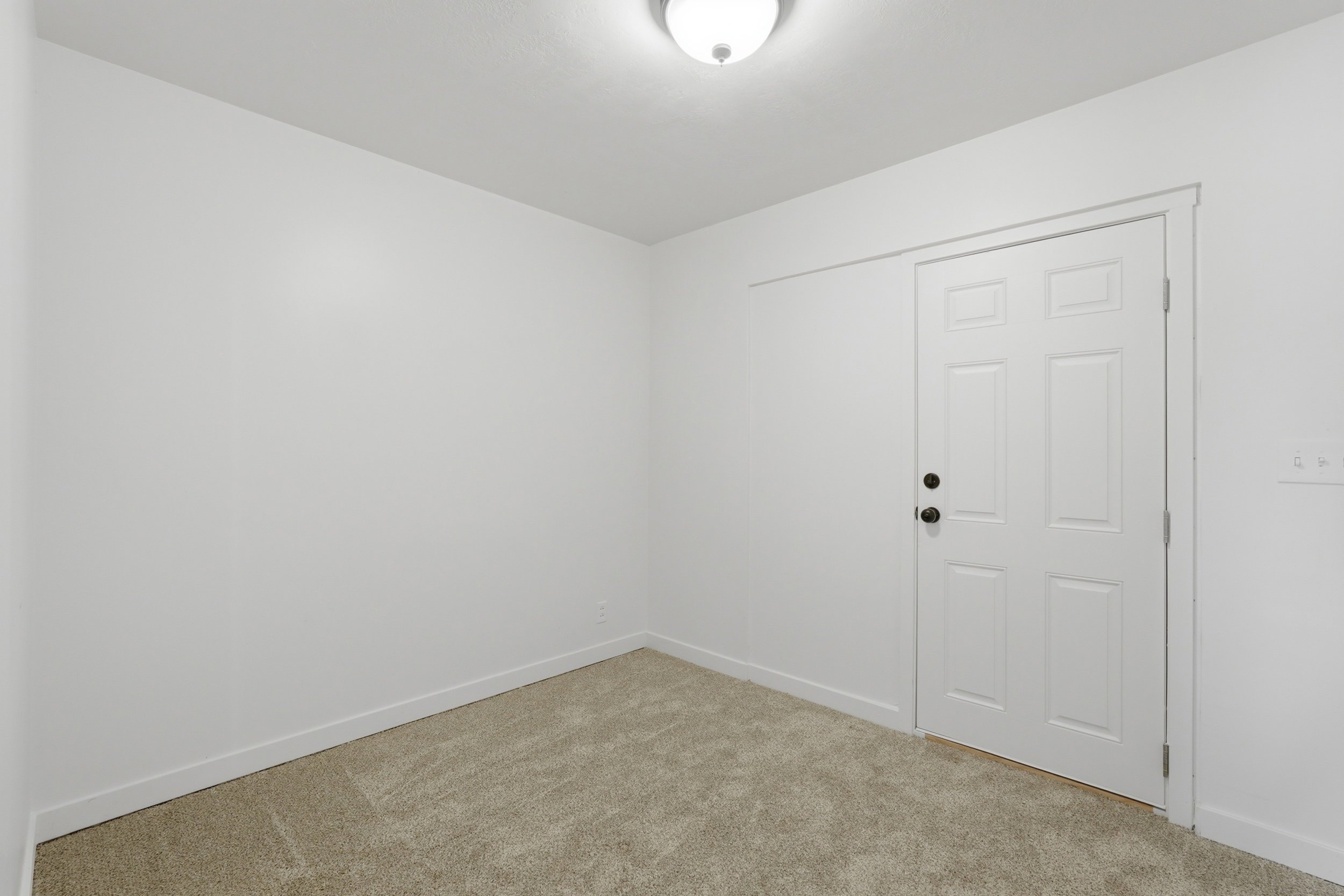 Room featuring light-toned carpeting, white walls, and a white six-panel door with matte black hardware