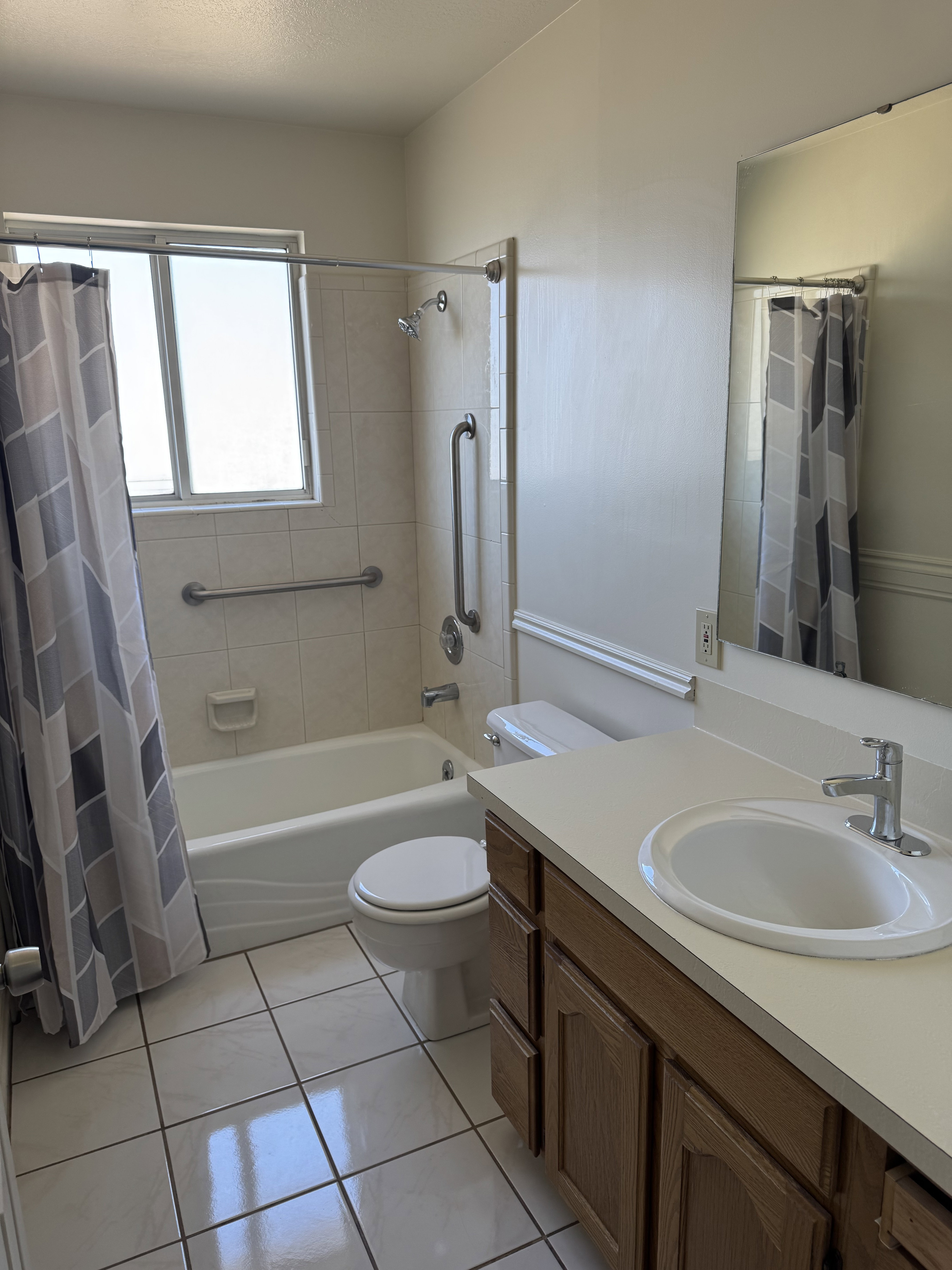 Bathroom featuring a white bathtub with grab bars, a window, a single-sink vanity with wood-finish cabinetry, a toilet, and tiled flooring