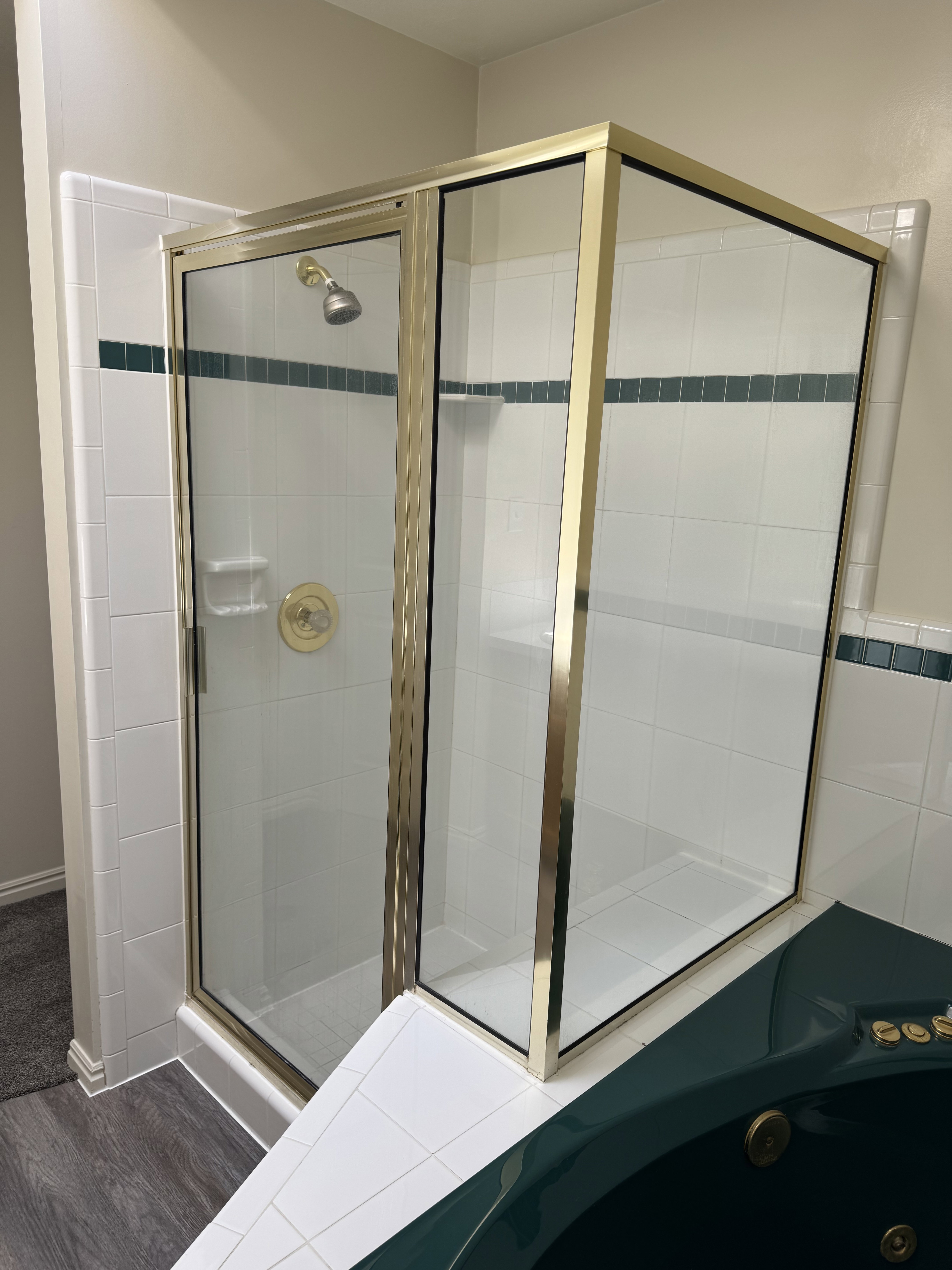 Gold-framed glass shower enclosure with a built-in shelf, white tile walls, a green accent tile band, and a coordinating green corner tub with brass fixtures