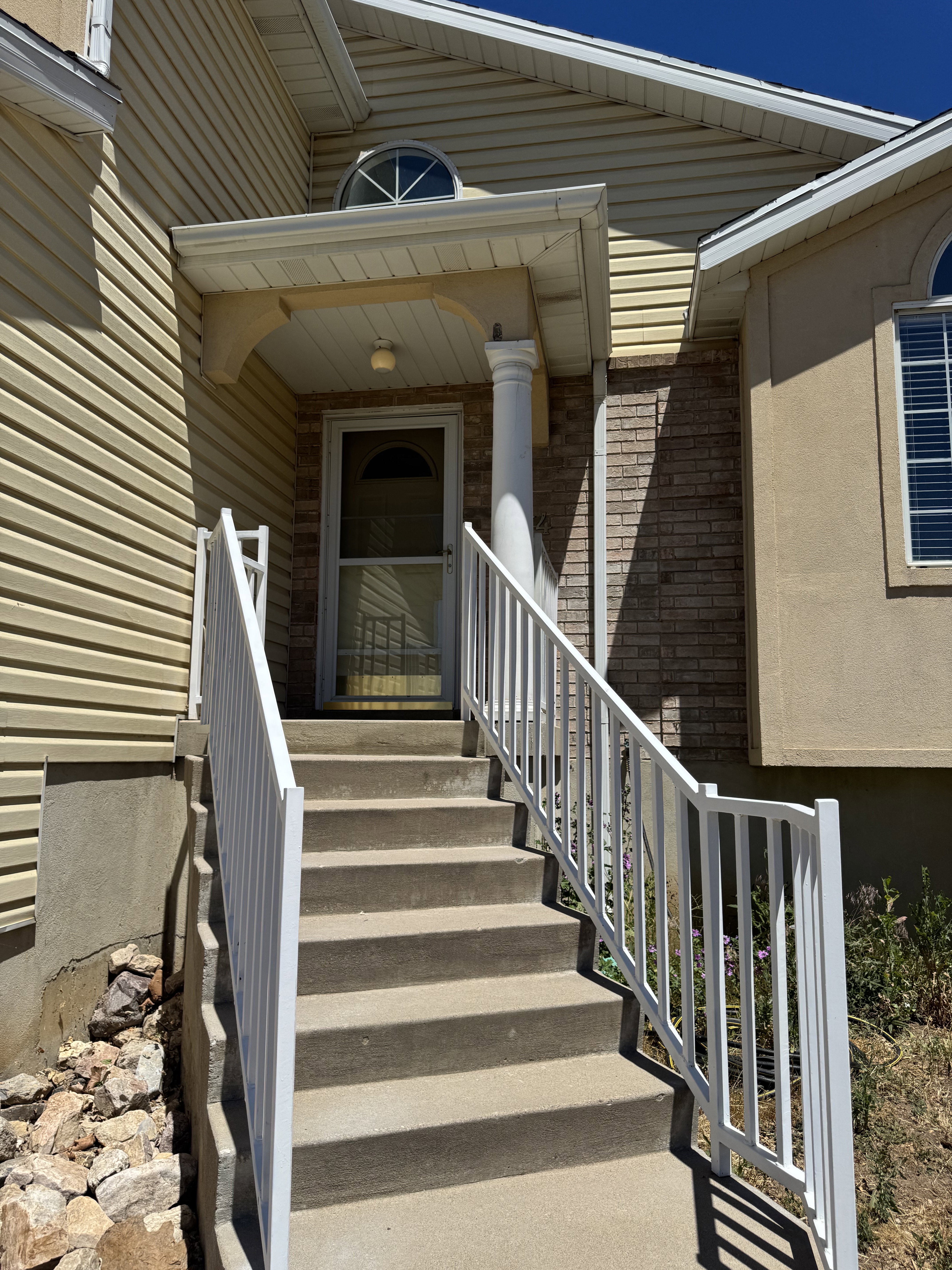 Inviting entryway featuring a covered porch, durable concrete steps, white metal railings, a half-round transom window, and a combination of siding and brick facade