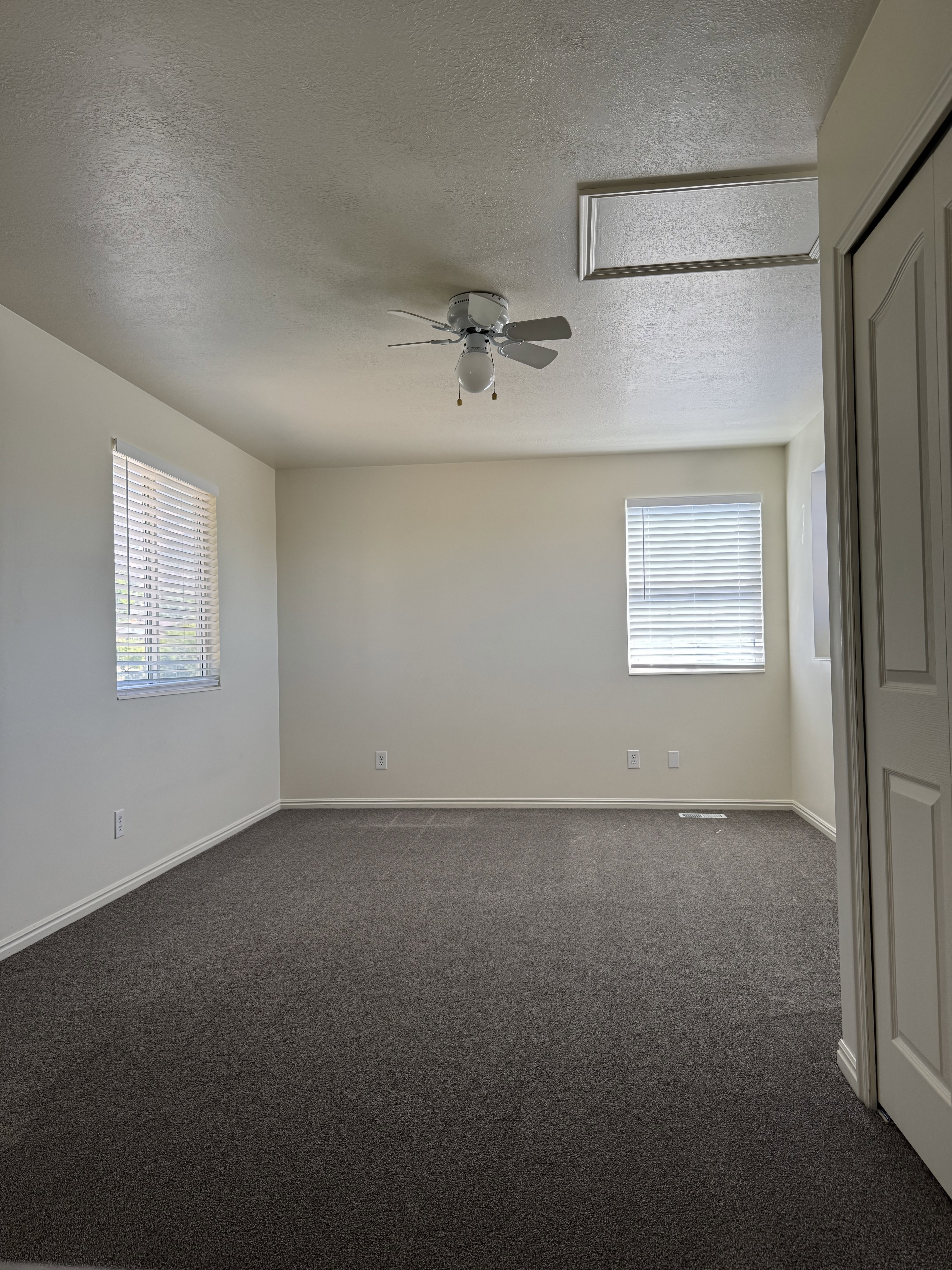 Spacious room featuring neutral carpeting, two windows with blinds, a ceiling fan with integrated lighting, and a white bi-fold closet door