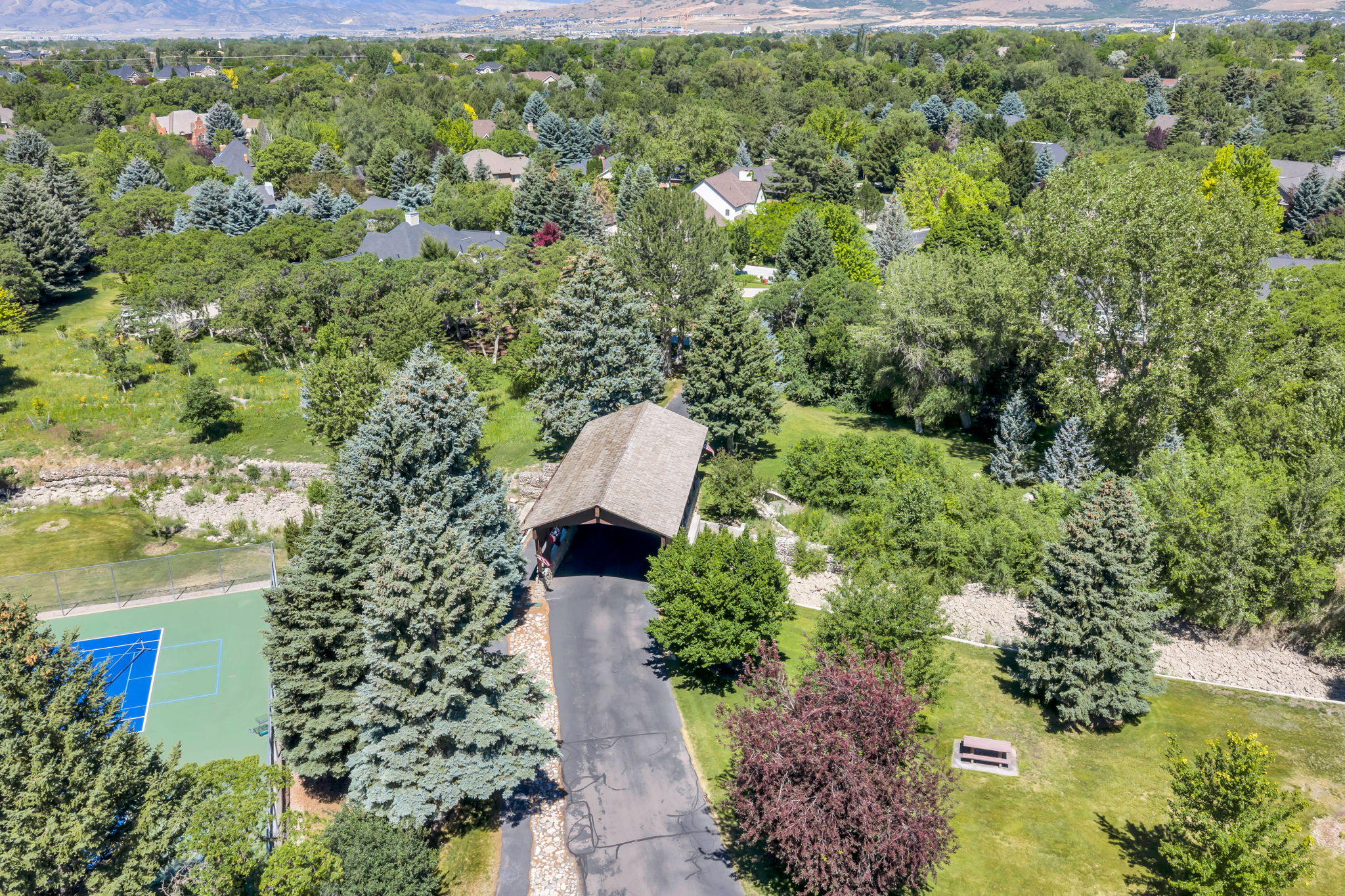 Expansive property featuring a private tennis court, covered parking structure, paved driveway, mature trees, and a mountain backdrop