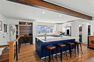 Open-concept kitchen featuring an exposed structural beam, white cabinetry, a large center island with blue base cabinetry, light-toned wood-finish flooring, and a window with outdoor tree scenery
