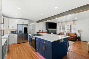 Open-concept kitchen and living area featuring wood-finish flooring, a stainless steel refrigerator, white upper and lower cabinetry, and a large central island with a built-in range