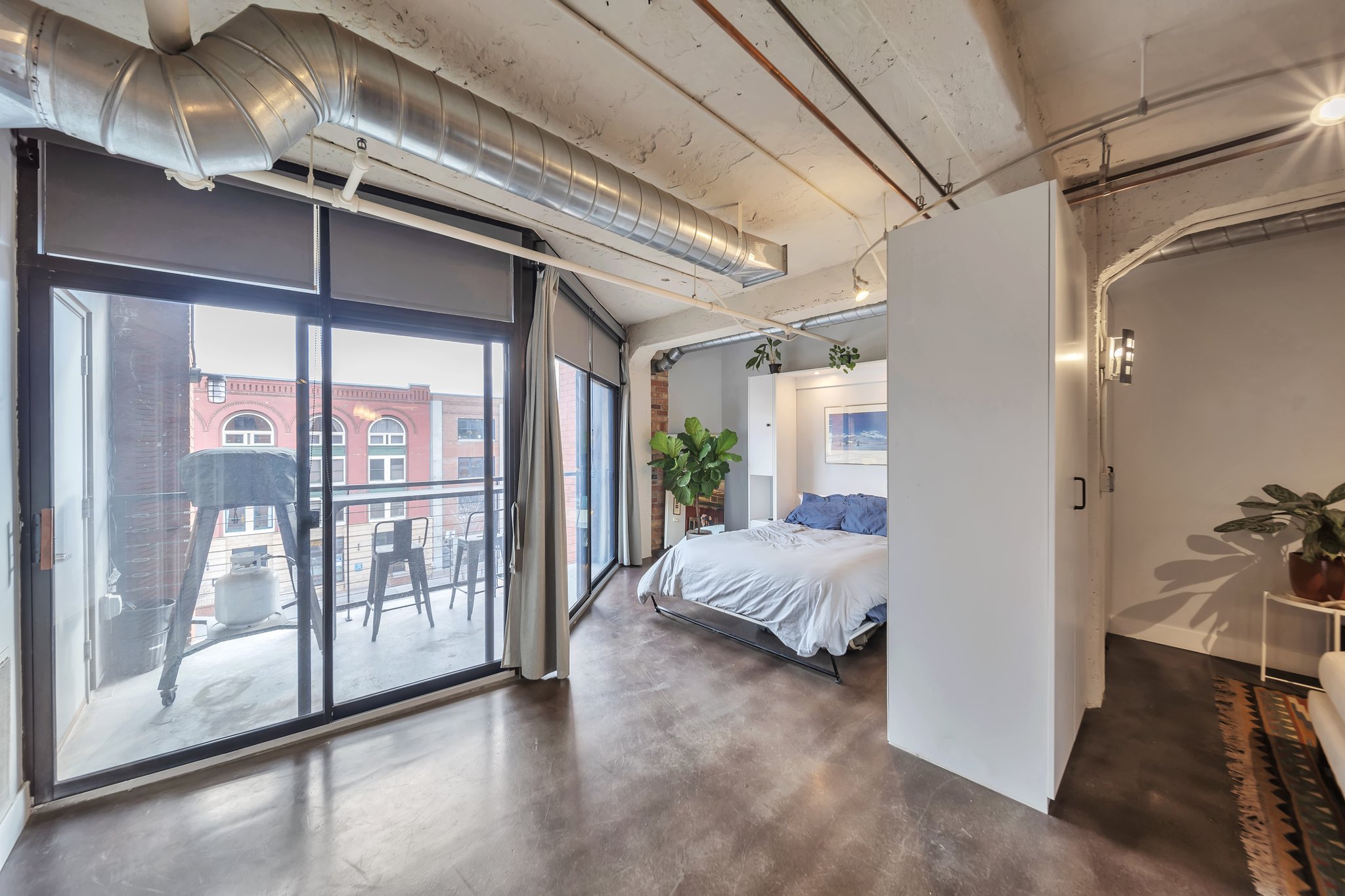 Spacious interior featuring a full-width sliding glass door to the balcony, exposed ductwork, concrete flooring, and a partial wall dividing the space