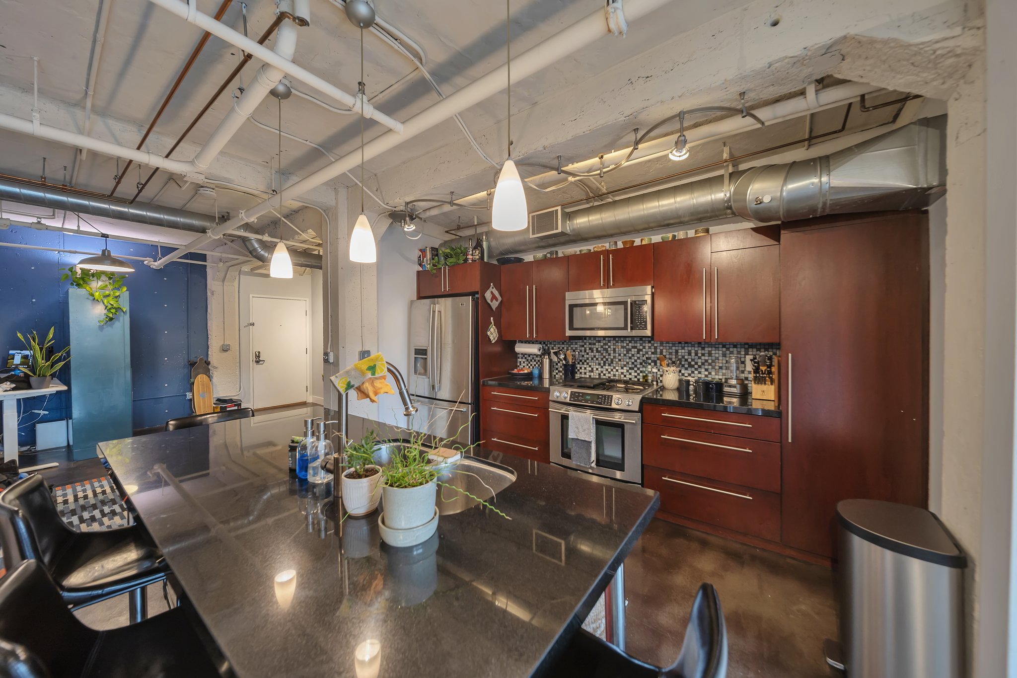 Modern kitchen featuring a large black countertop island with integrated sink, dark wood-finish cabinetry, stainless steel appliances, and a mosaic tile backsplash