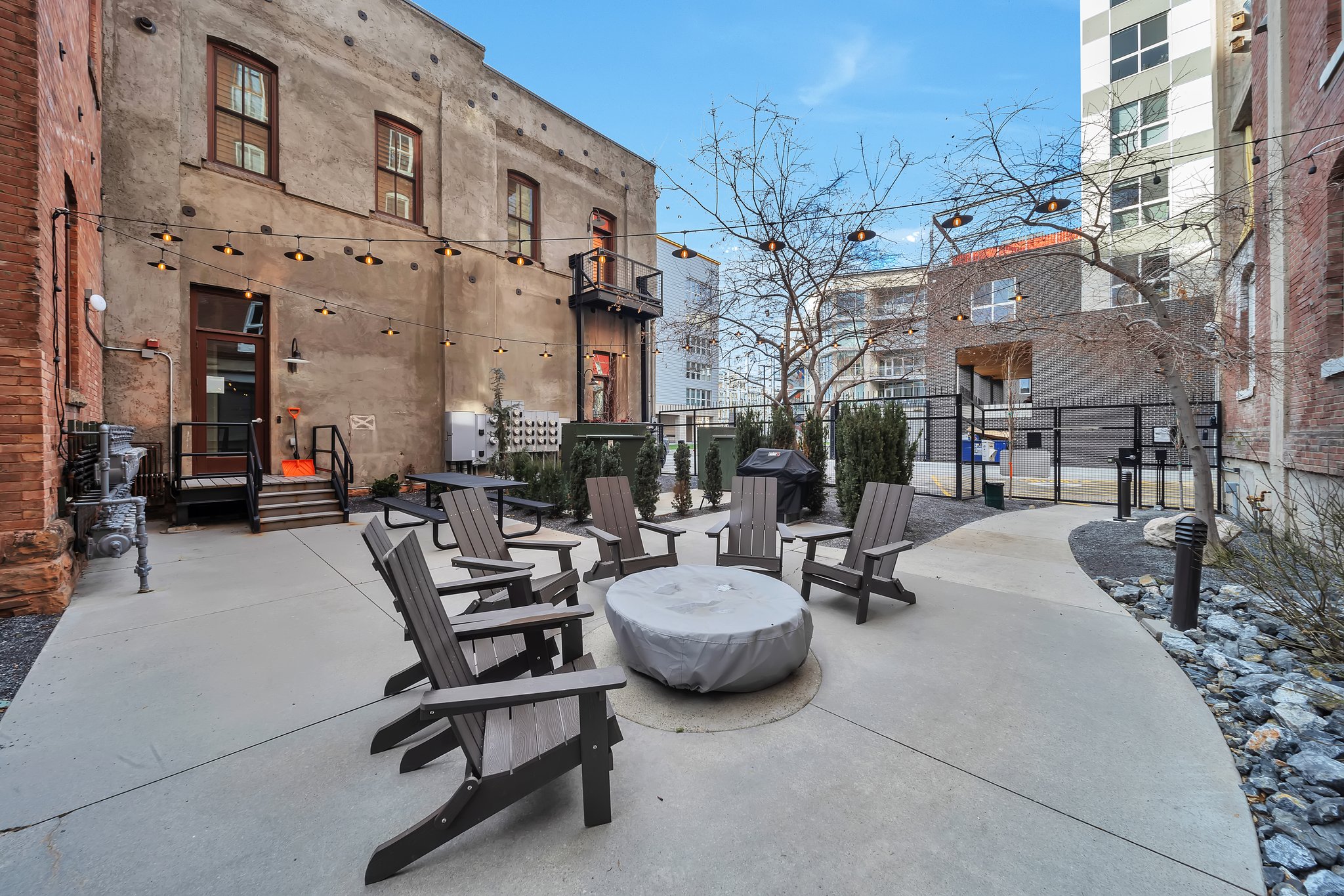 Expansive concrete patio featuring a central fire pit, surrounded by Adirondack chairs, string lighting, and a picnic table