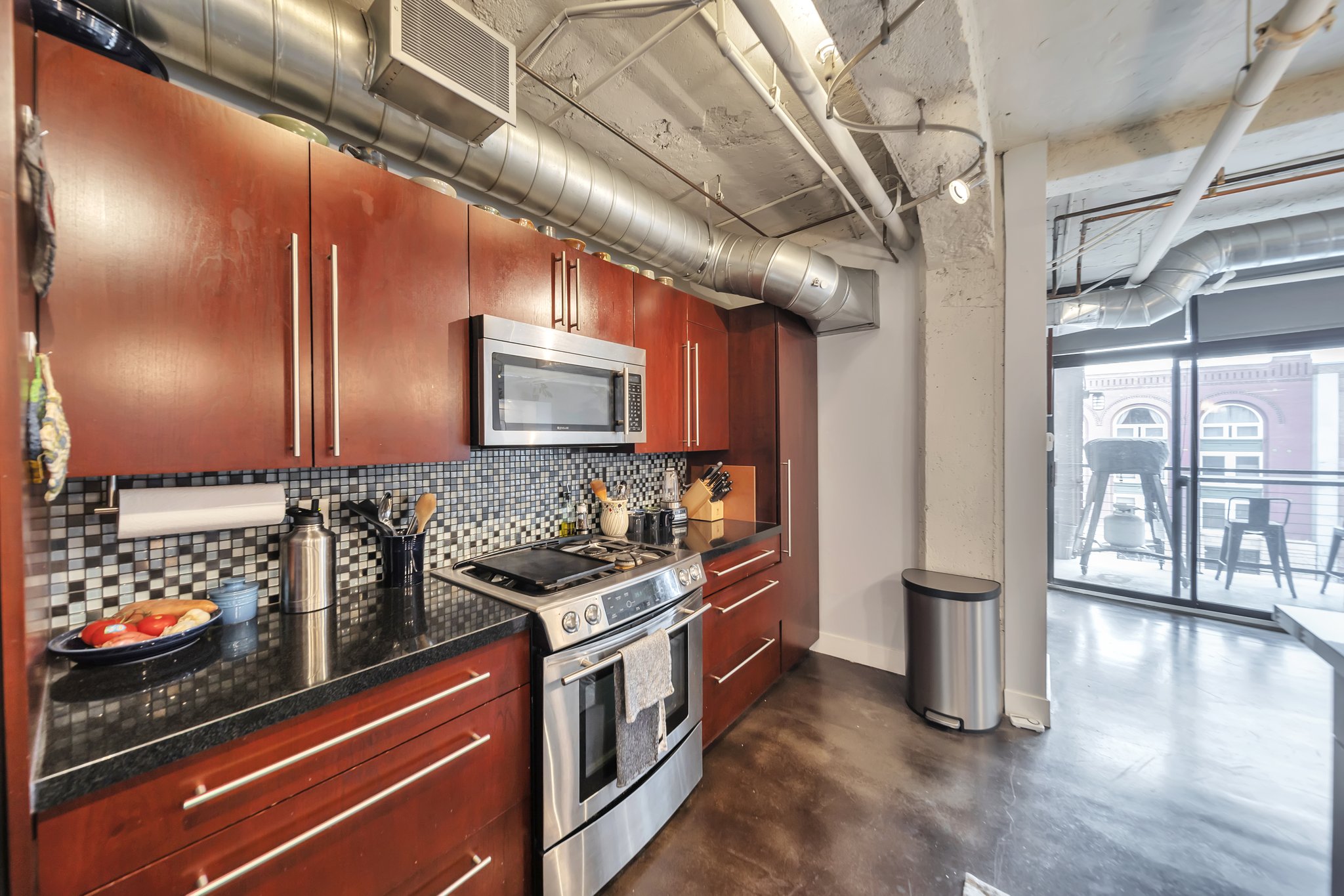 Modern kitchen featuring wood-finish cabinetry, stainless steel appliances, dark countertops, and a mosaic tile backsplash