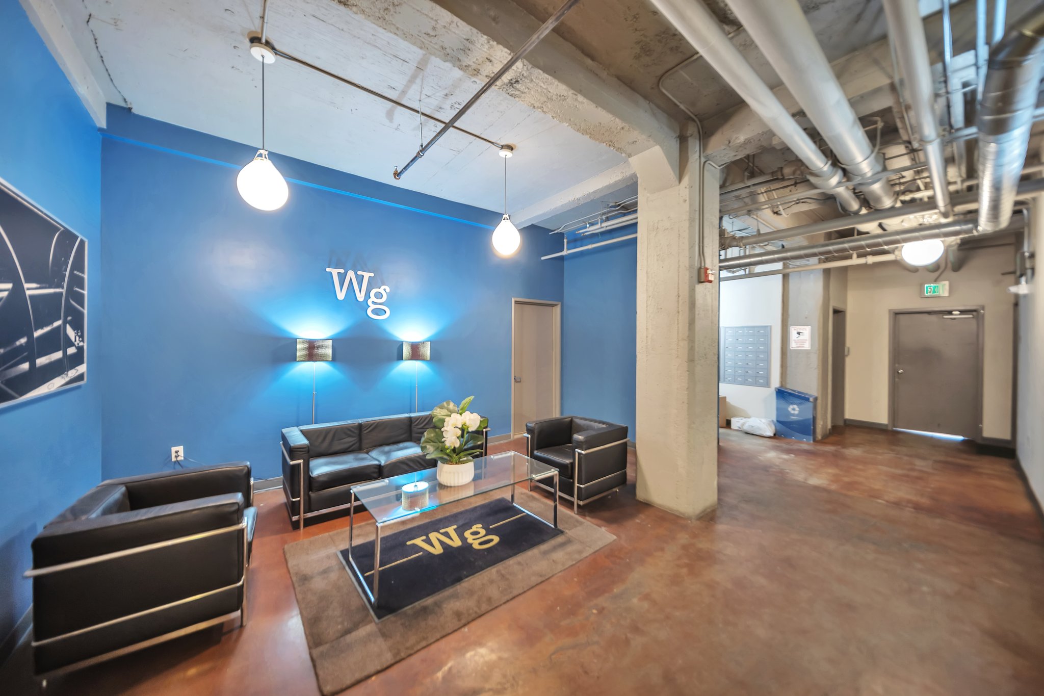 Lobby area featuring a blue accent wall with ambient sconce lighting, suspended pendant lights, exposed industrial-style ceiling ductwork, and concrete flooring